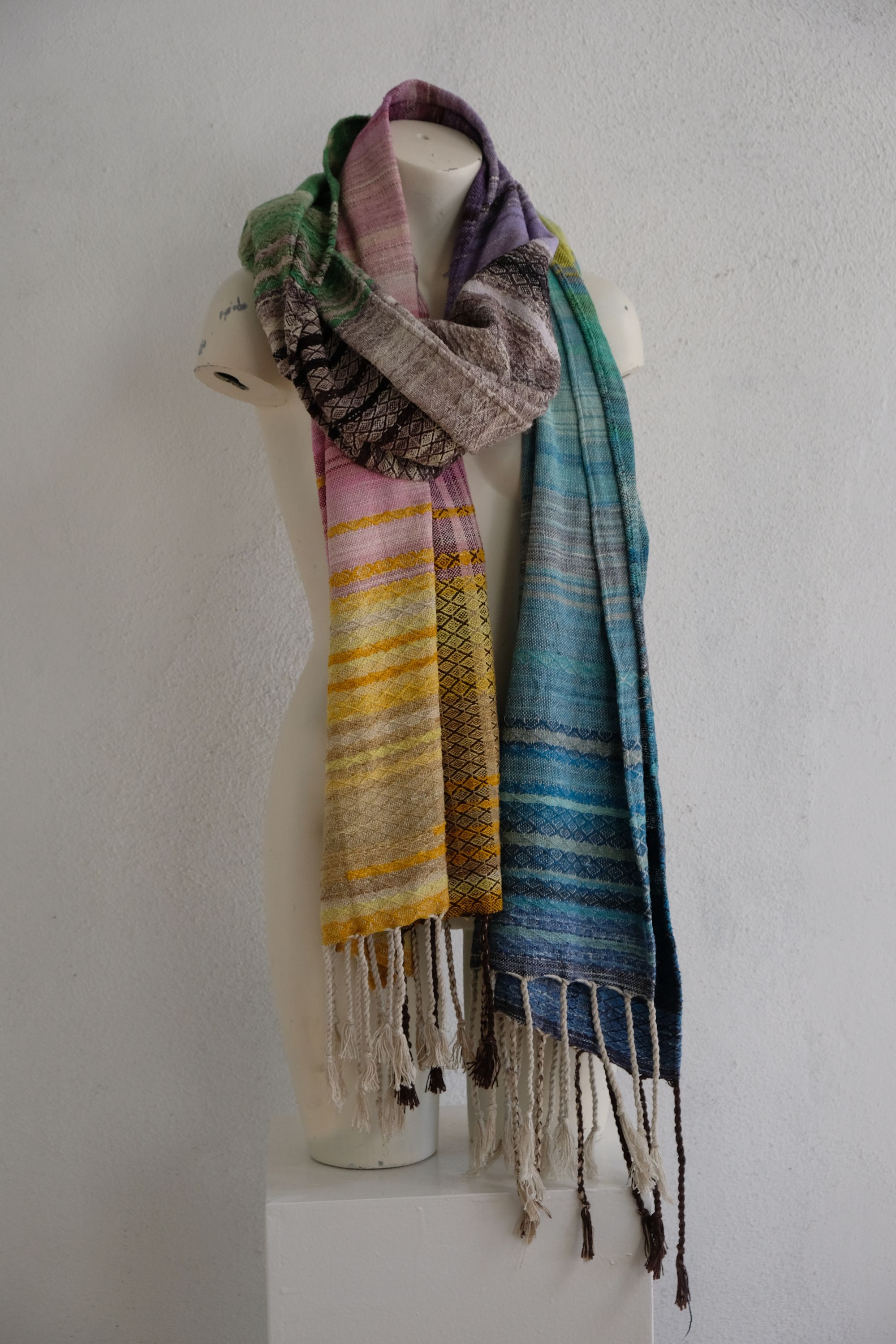 handwoven, hand dyed in blues, green, gray, black, purple, pink and yellow,  fabric of many colors with a diamond pattern, is draped on a mannequin in a white gallery space