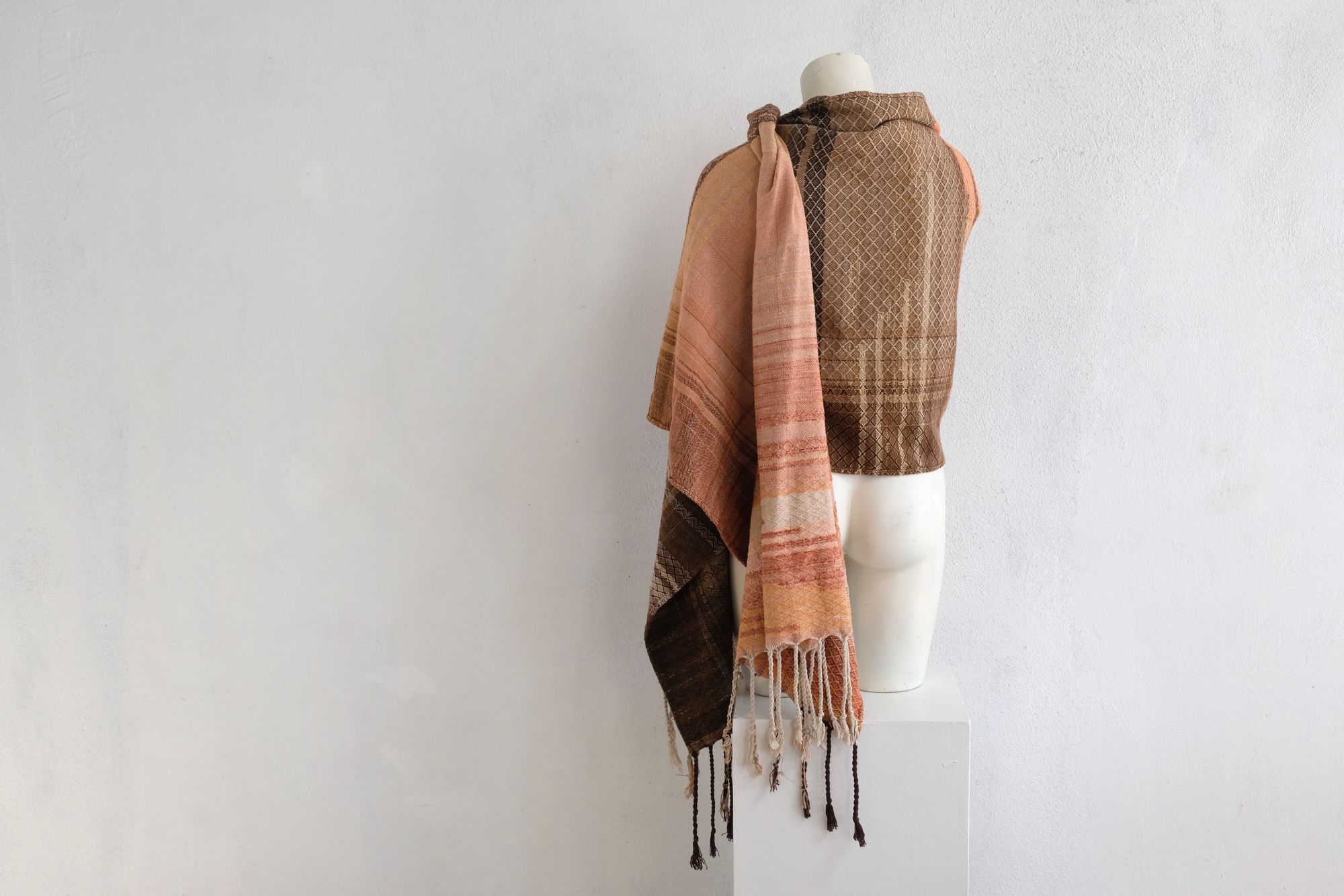 A handwoven shawl is draped on a white painted mannequin against a white wall. The shawl is naturally dyed in shades of earthy browns, oranges and peach pinks