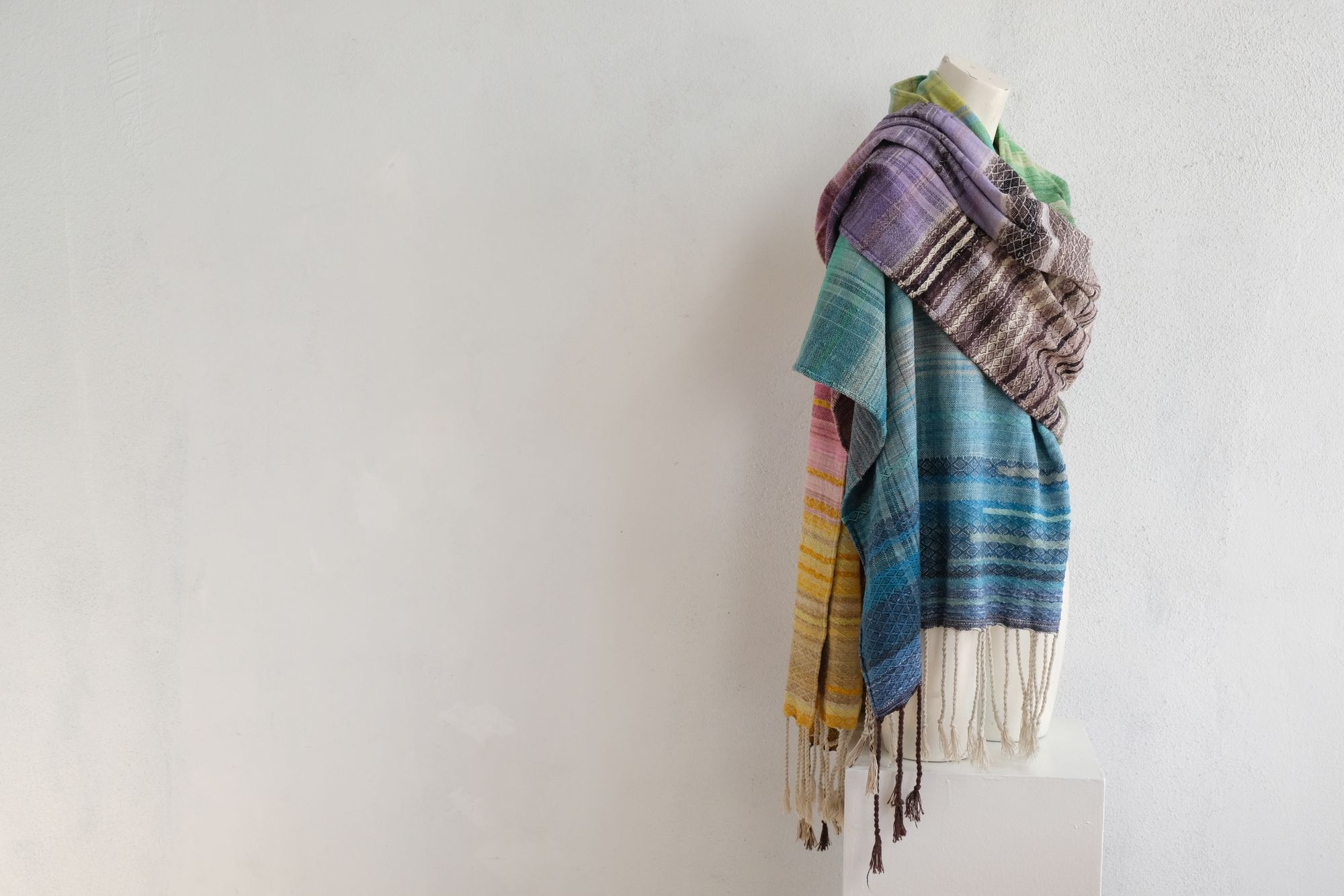 handwoven, hand dyed in blues, green, gray, black, purple, pink and yellow,  fabric of many colors with a diamond pattern, is draped on a mannequin in a white gallery space