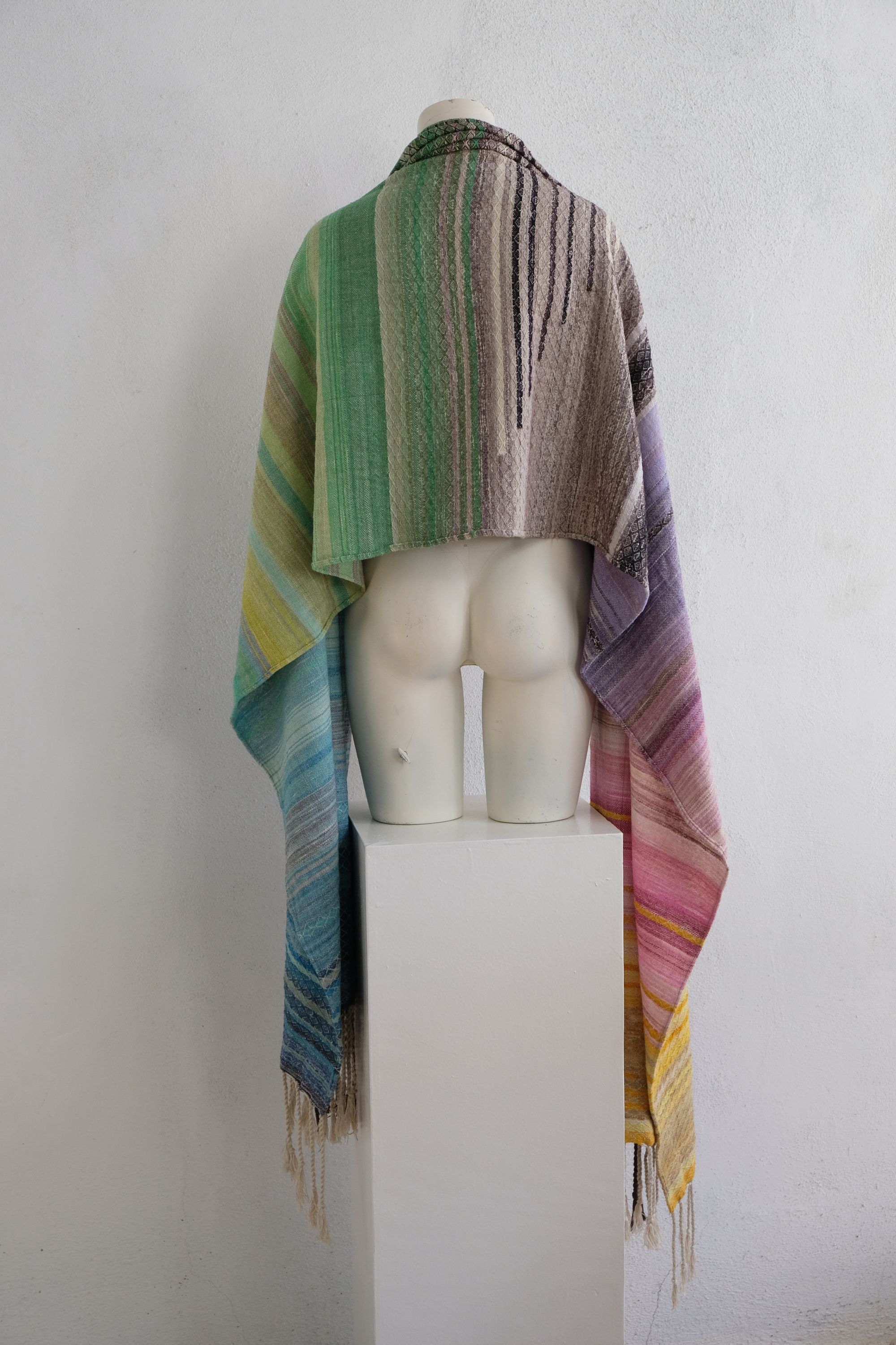 handwoven, hand dyed in blues, green, gray, black, purple, pink and yellow,  fabric of many colors with a diamond pattern, is draped on a mannequin in a white gallery space