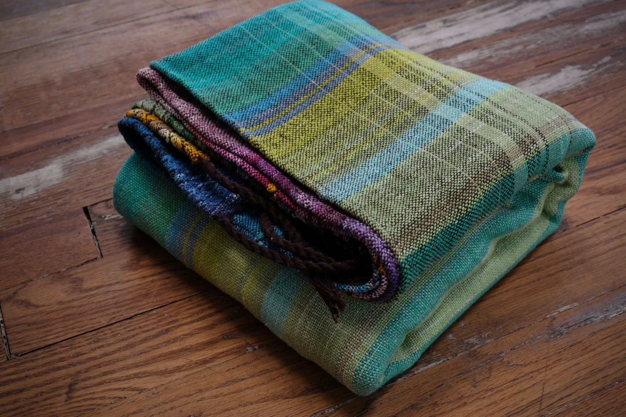 handwoven, hand dyed fabric of many colors with a diamond pattern, folded on a wooden floor.