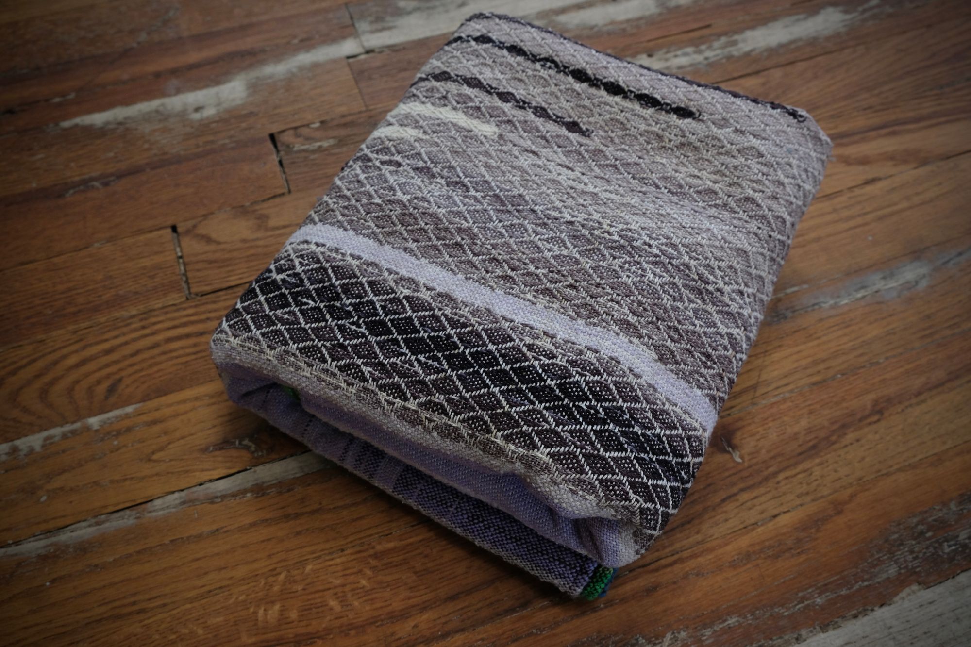 handwoven, hand dyed fabric of many colors with a diamond pattern, folded on a wooden floor.