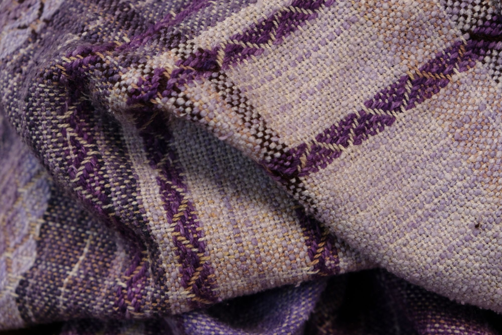 A handwoven length of fabric, woven in every shade of purple with a diamond texture pattern, is laid out on a wooden floor.