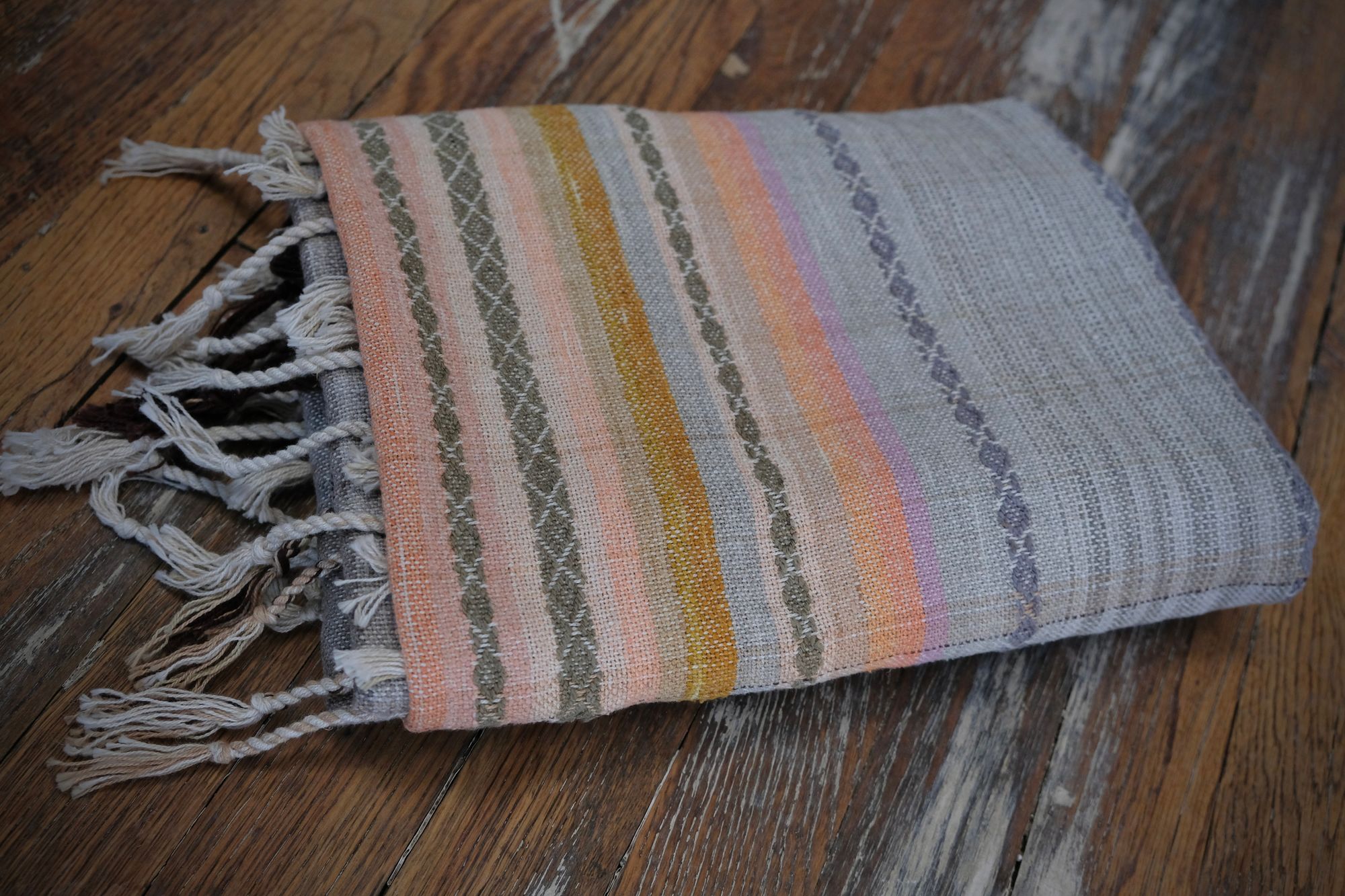 Details of a handwoven shawl in soft but brilliant naturally dyed shades of gray, yellow, pink, salmon, orange, green and purple. The shawl is designed with small stripes of all these colors, and pattern-texture woven details, also of tiny stripes