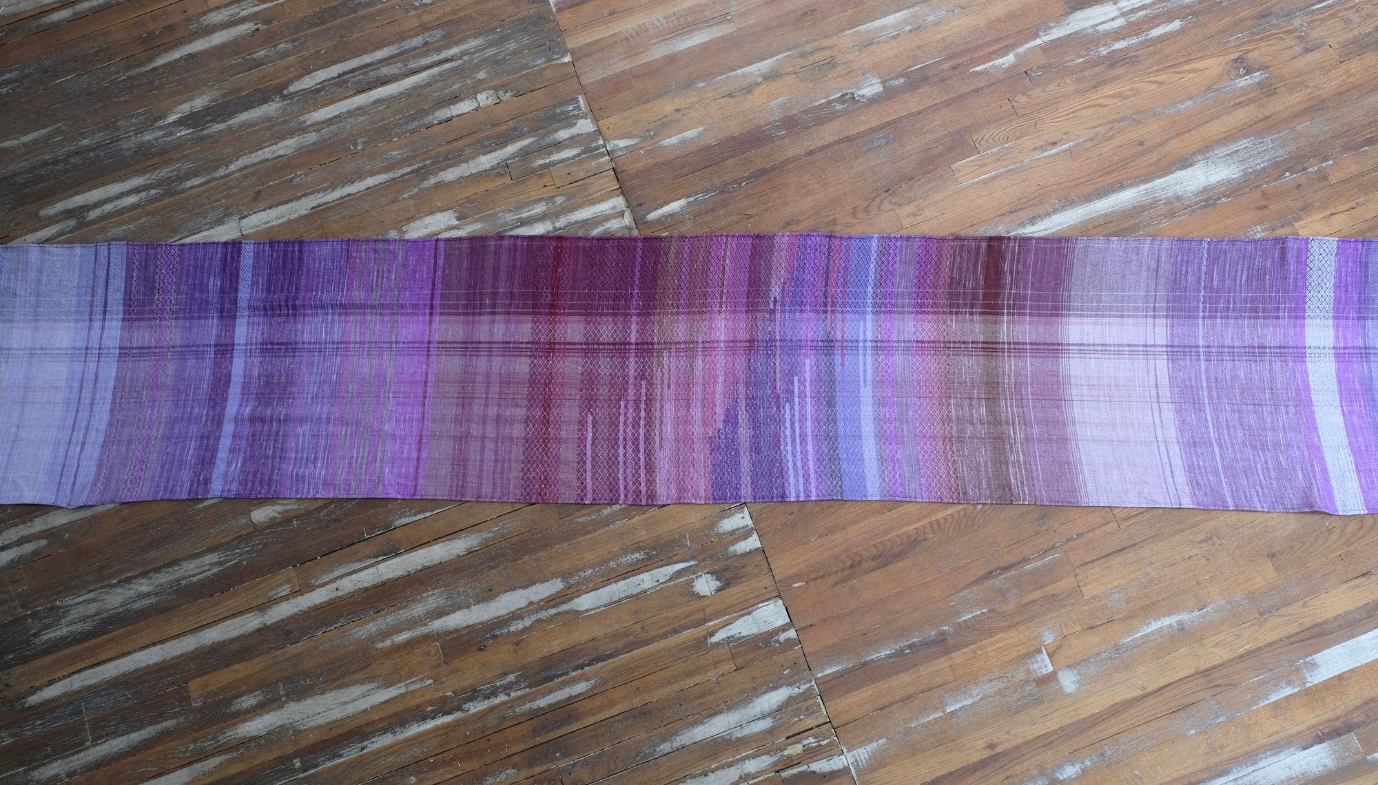 A handwoven length of fabric, woven in every shade of purple is laid out on a wooden floor