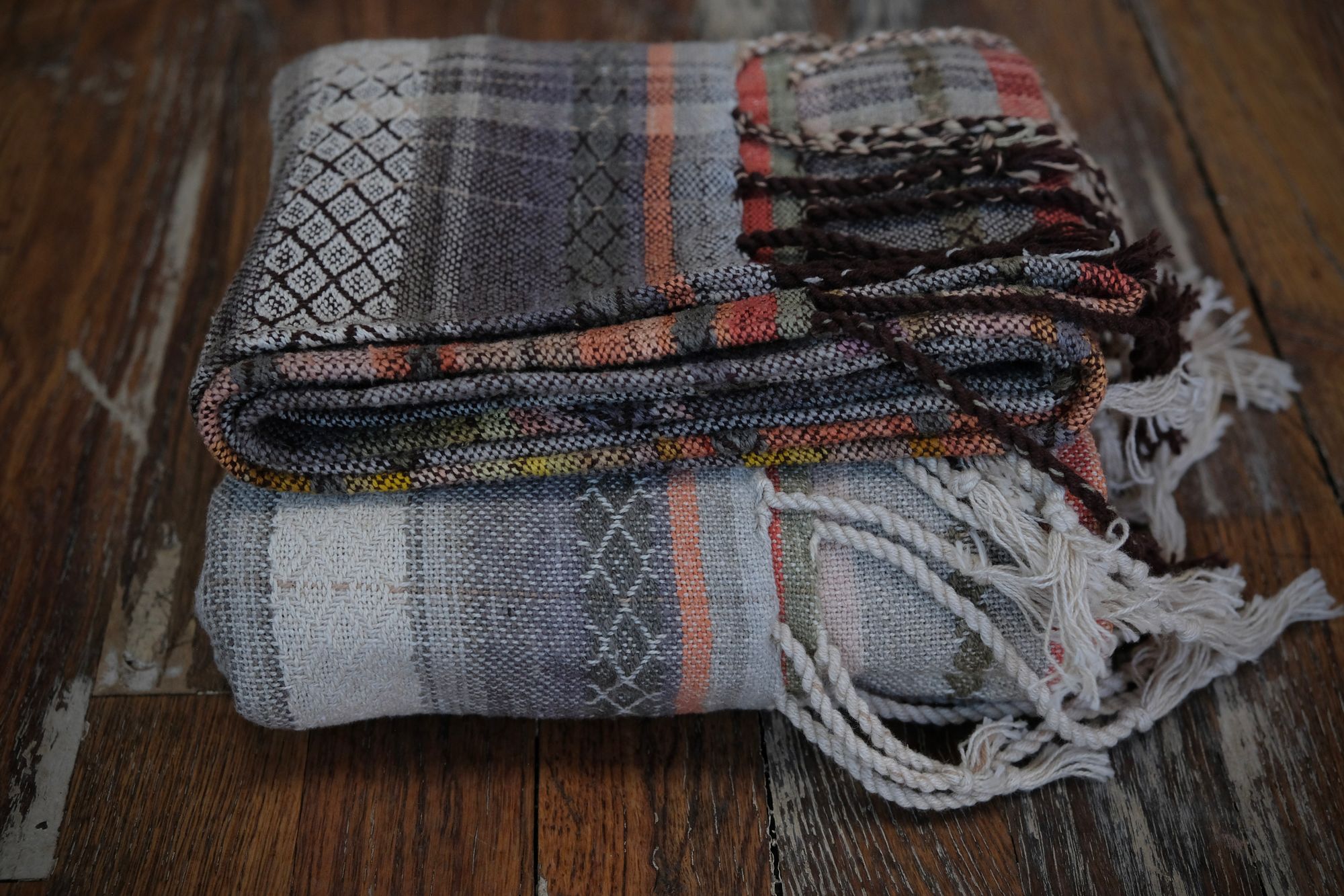 Details of a handwoven shawl in soft but brilliant naturally dyed shades of gray, yellow, pink, salmon, orange, green and purple. The shawl is designed with small stripes of all these colors, and pattern-texture woven details, also of tiny stripes. The shawl is folded on a wooden floor