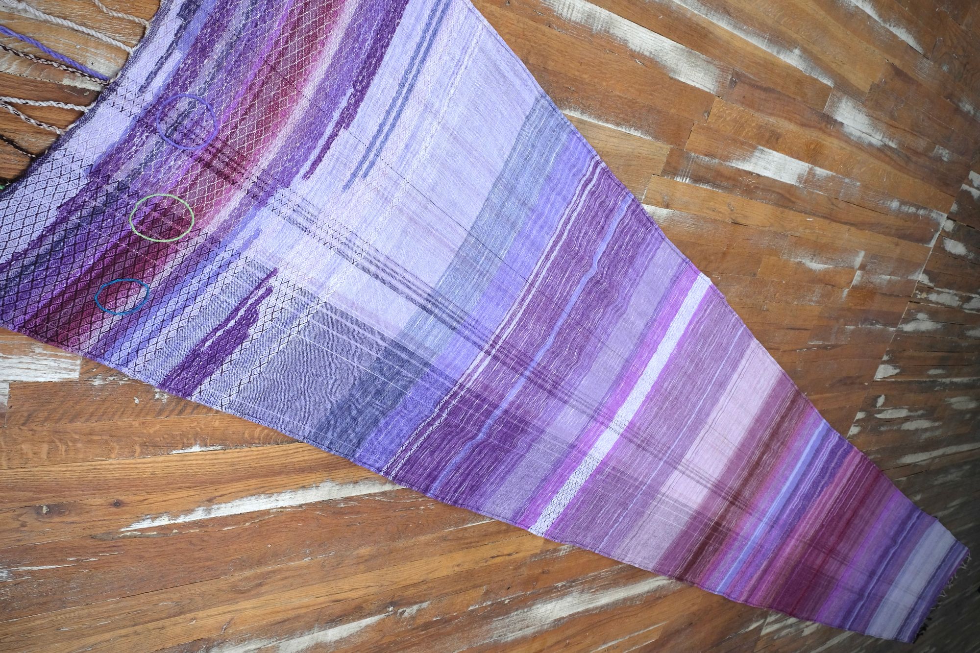 A handwoven length of fabric, woven in every shade of purple is laid out on a wooden floor. It has details of three circles, one sapphire blue, one peridot green and one tanzanite purple