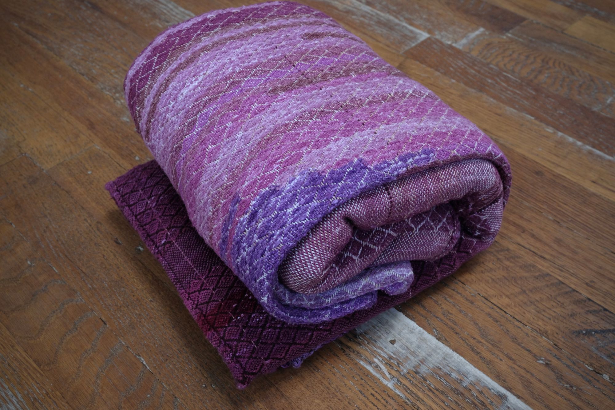 A handwoven length of fabric, woven in every shade of purple with a diamond texture pattern, is folded on a wooden floor.