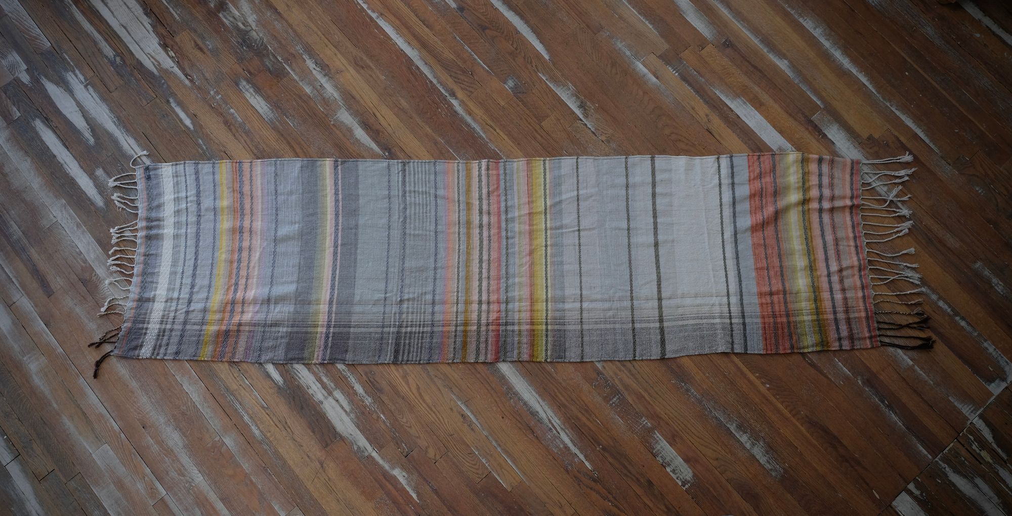 Details of a handwoven shawl in soft but brilliant naturally dyed shades of gray, yellow, pink, salmon, orange, green and purple. The shawl is designed with small stripes of all these colors, and pattern-texture woven details, also of tiny stripes. The shawl is laid flat onto a wooden floor