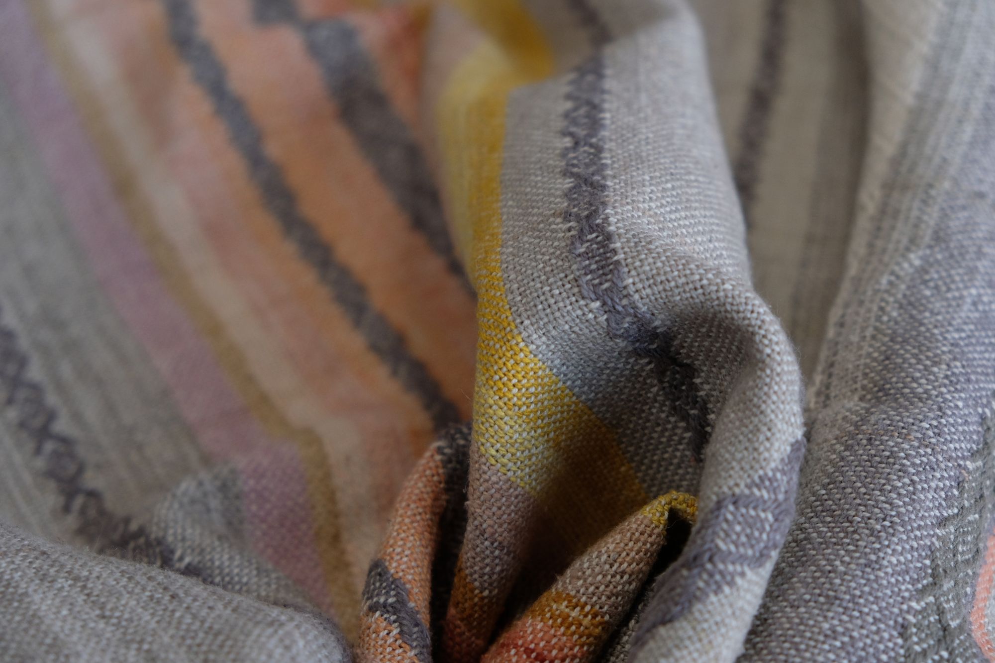 Details of a handwoven shawl in soft but brilliant naturally dyed shades of gray, yellow, pink, salmon, orange, green and purple. The shawl is designed with small stripes of all these colors, and pattern-texture woven details, also of tiny stripes