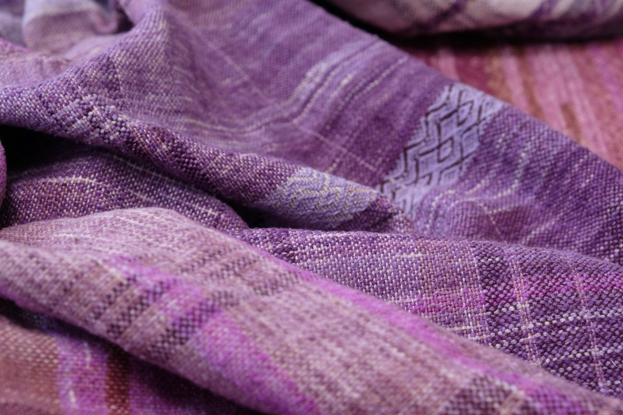 A handwoven length of fabric, woven in every shade of purple is laid out on a wooden floor. It has details of three circles, one sapphire blue, one peridot green and one tanzanite purple