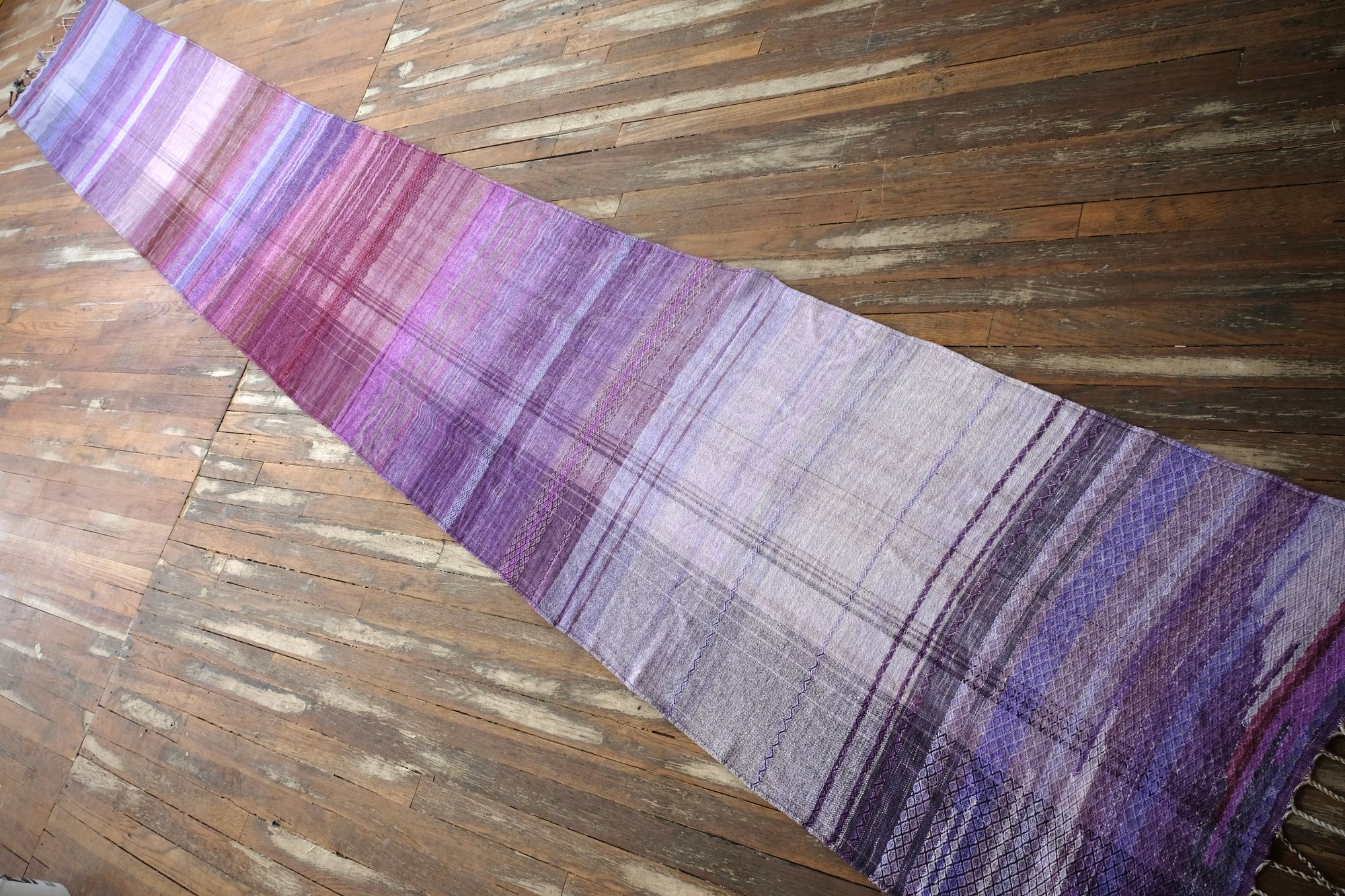 A handwoven length of fabric, woven in every shade of purple is laid out on a wooden floor. It has details of three circles, one sapphire blue, one peridot green and one tanzanite purple