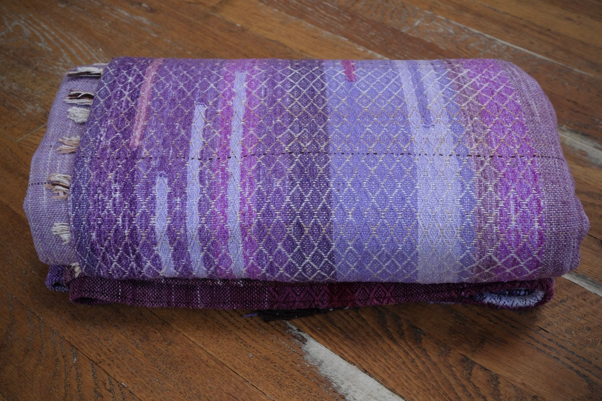 A handwoven length of fabric, woven in every shade of purple with a diamond texture pattern, is folded on a wooden floor.