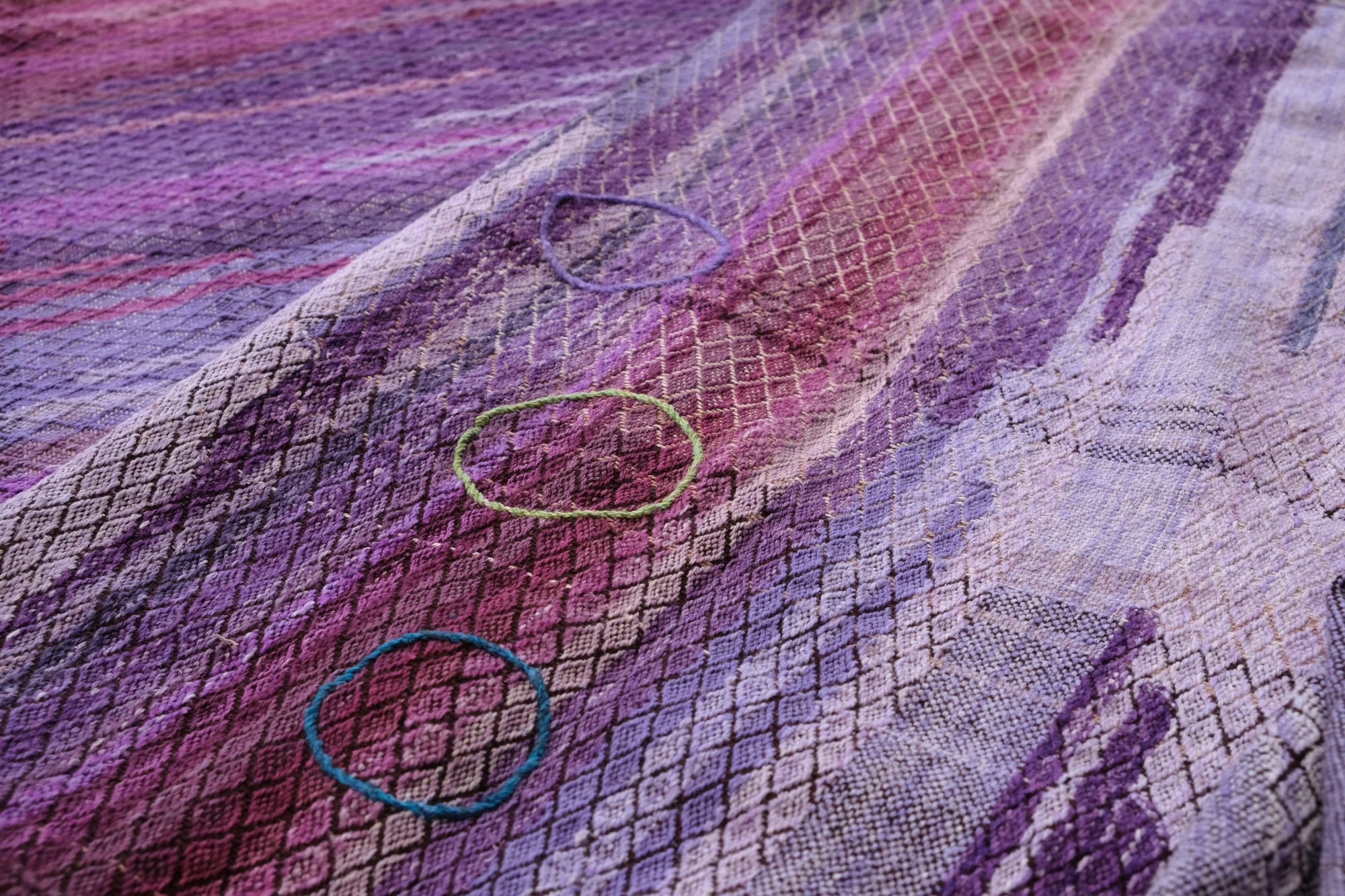 A handwoven length of fabric, woven in every shade of purple is laid out on a wooden floor. It has details of three circles, one sapphire blue, one peridot green and one tanzanite purple