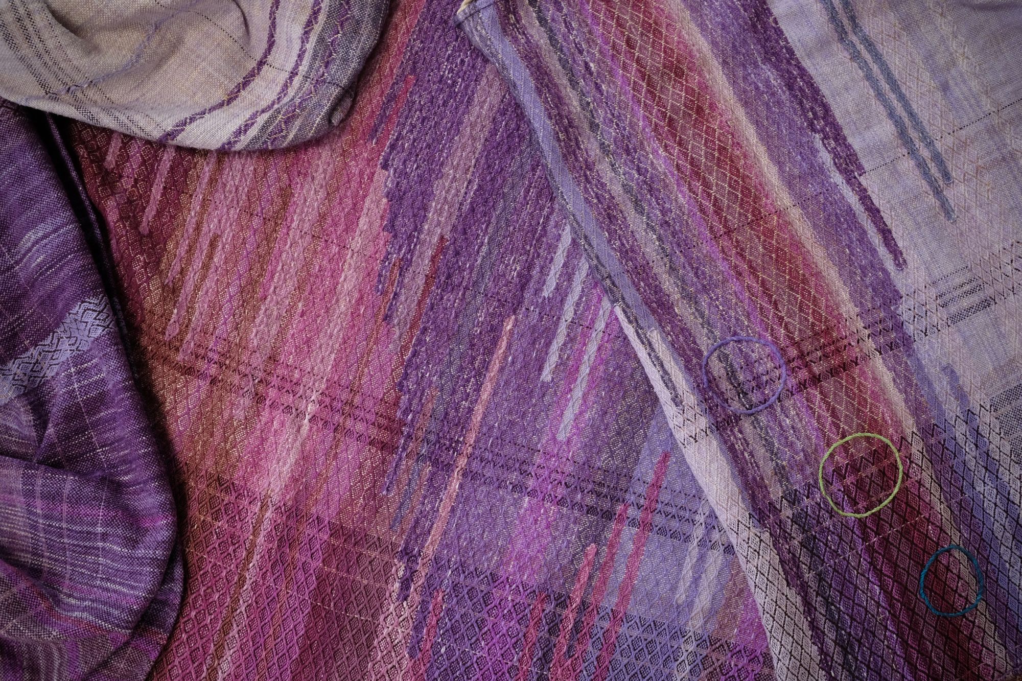 A handwoven length of fabric, woven in every shade of purple is laid out on a wooden floor. It has details of three circles, one sapphire blue, one peridot green and one tanzanite purple