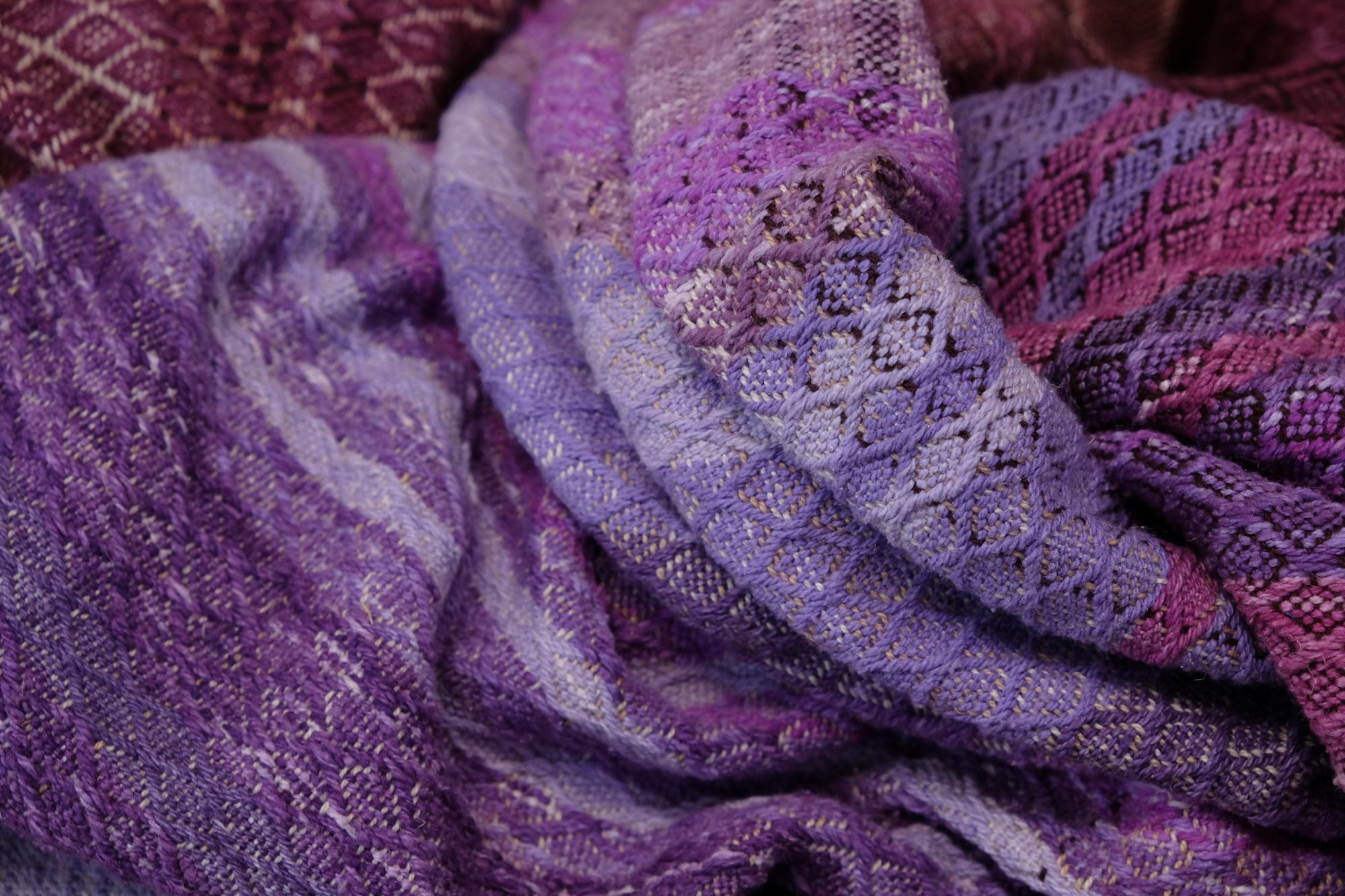 A handwoven length of fabric, woven in every shade of purple is laid out on a wooden floor. It has details of three circles, one sapphire blue, one peridot green and one tanzanite purpleA handwoven length of fabric, woven in every shade of purple with a diamond texture pattern, is laid out on a wooden floor.