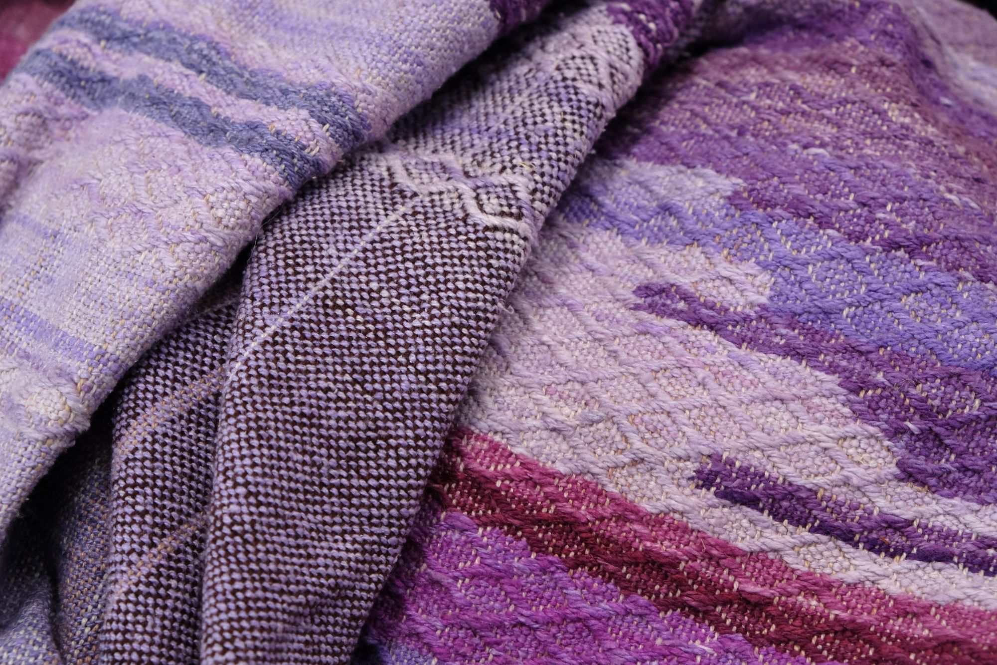 A handwoven length of fabric, woven in every shade of purple with a diamond texture pattern, is laid out on a wooden floor.