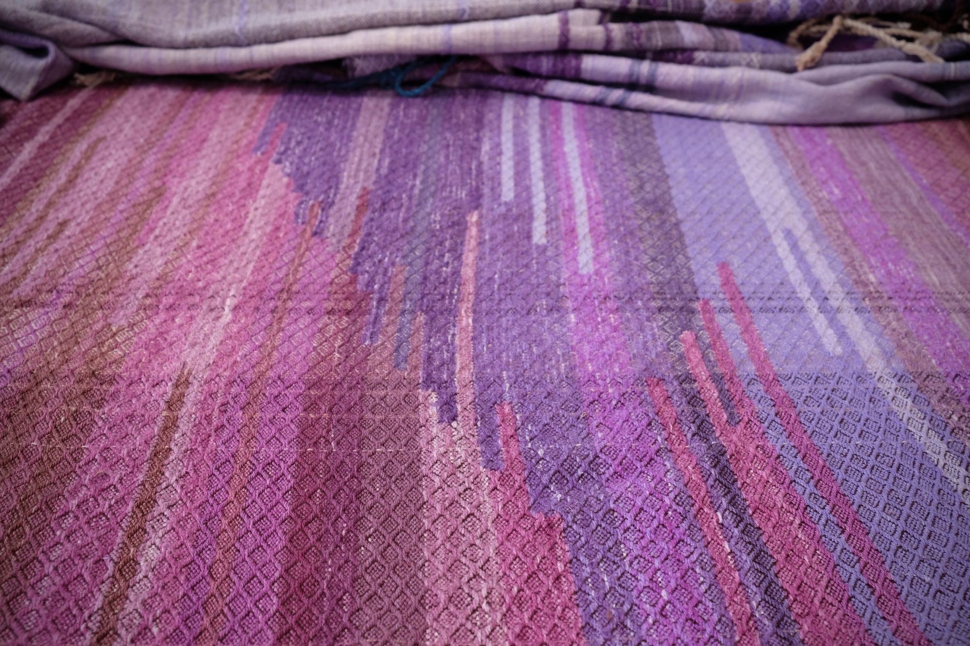 A handwoven length of fabric, woven in every shade of purple is laid out on a wooden floor