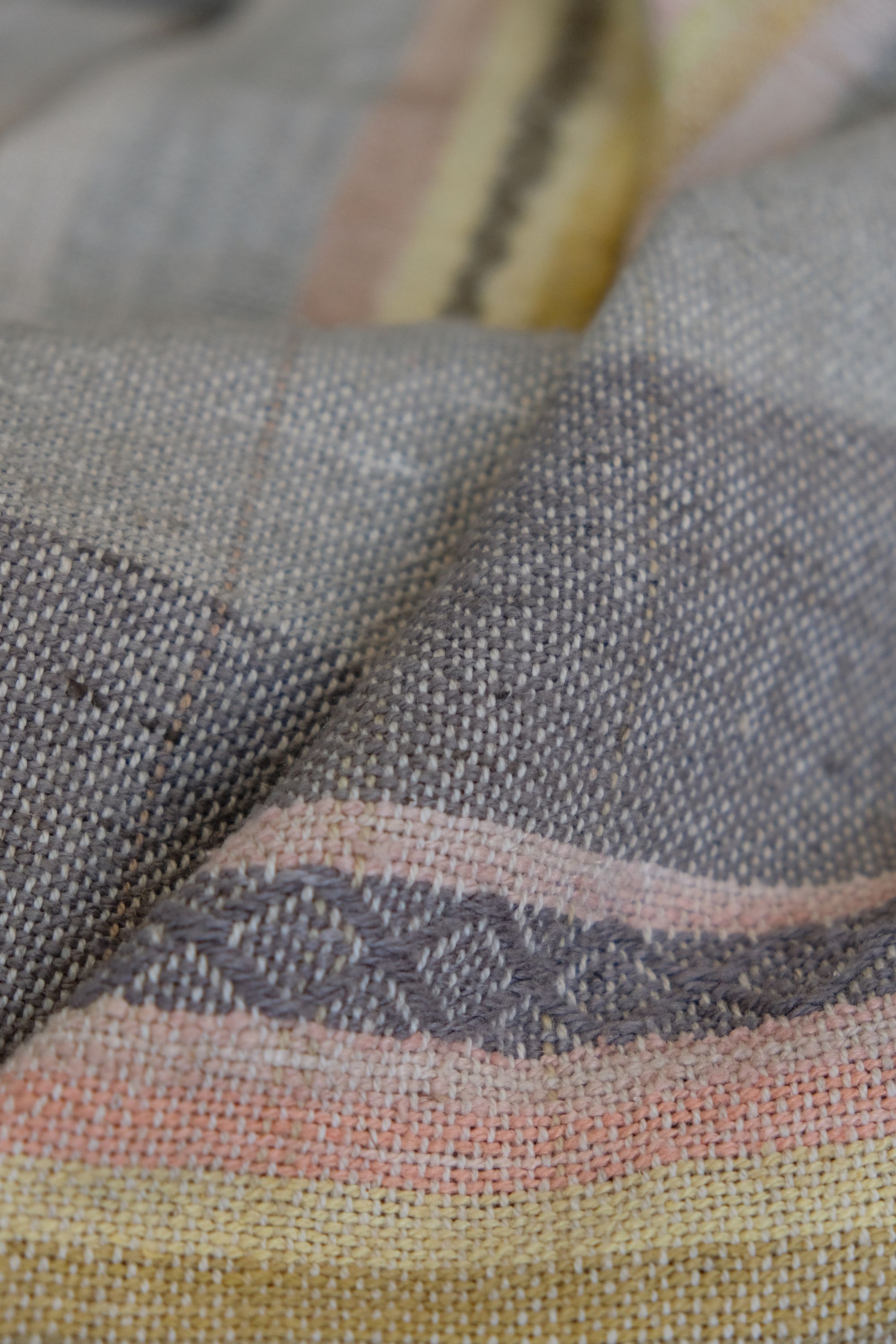 Details of a handwoven shawl in soft but brilliant naturally dyed shades of gray, yellow, pink, salmon, orange, green and purple. The shawl is designed with small stripes of all these colors, and pattern-texture woven details, also of tiny stripes