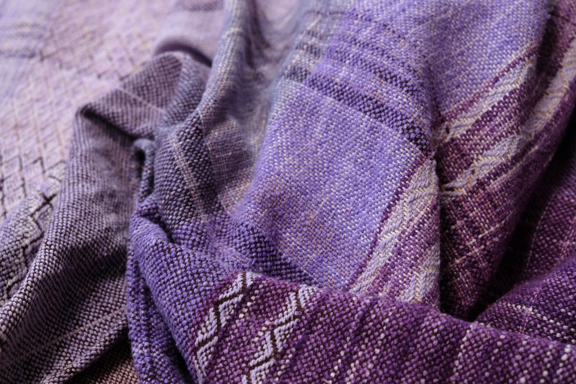 A handwoven length of fabric, woven in every shade of purple is laid out on a wooden floor. It has details of three circles, one sapphire blue, one peridot green and one tanzanite purple