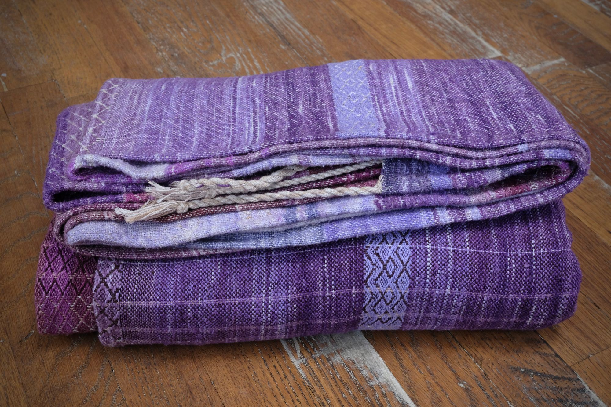 A handwoven length of fabric, woven in every shade of purple with a diamond texture pattern, is folded on a wooden floor.