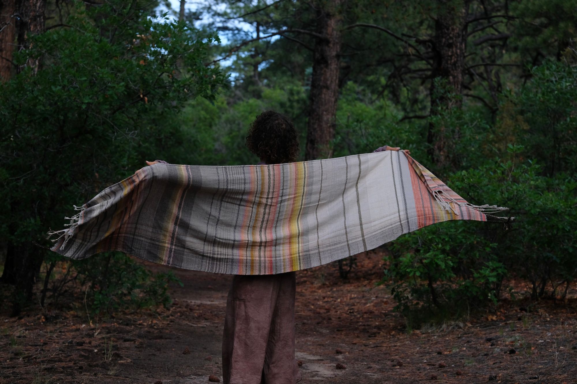 A woman stands on a trail in the forest wearing a handwoven shawl in subtle naturally dyed colors