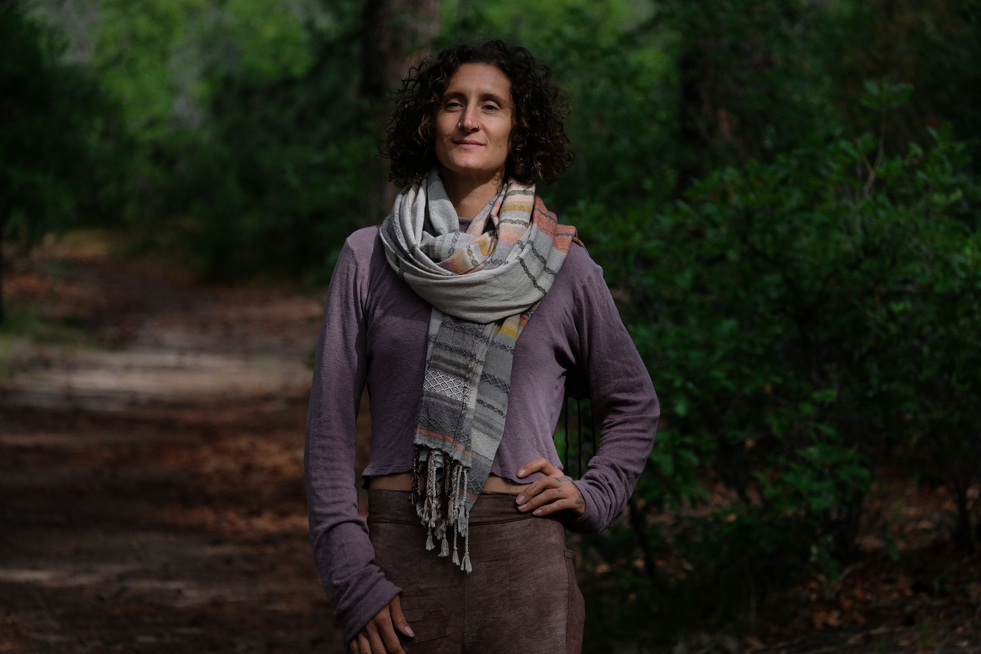 A woman stands on a trail in the forest wearing a handwoven shawl in subtle naturally dyed colors