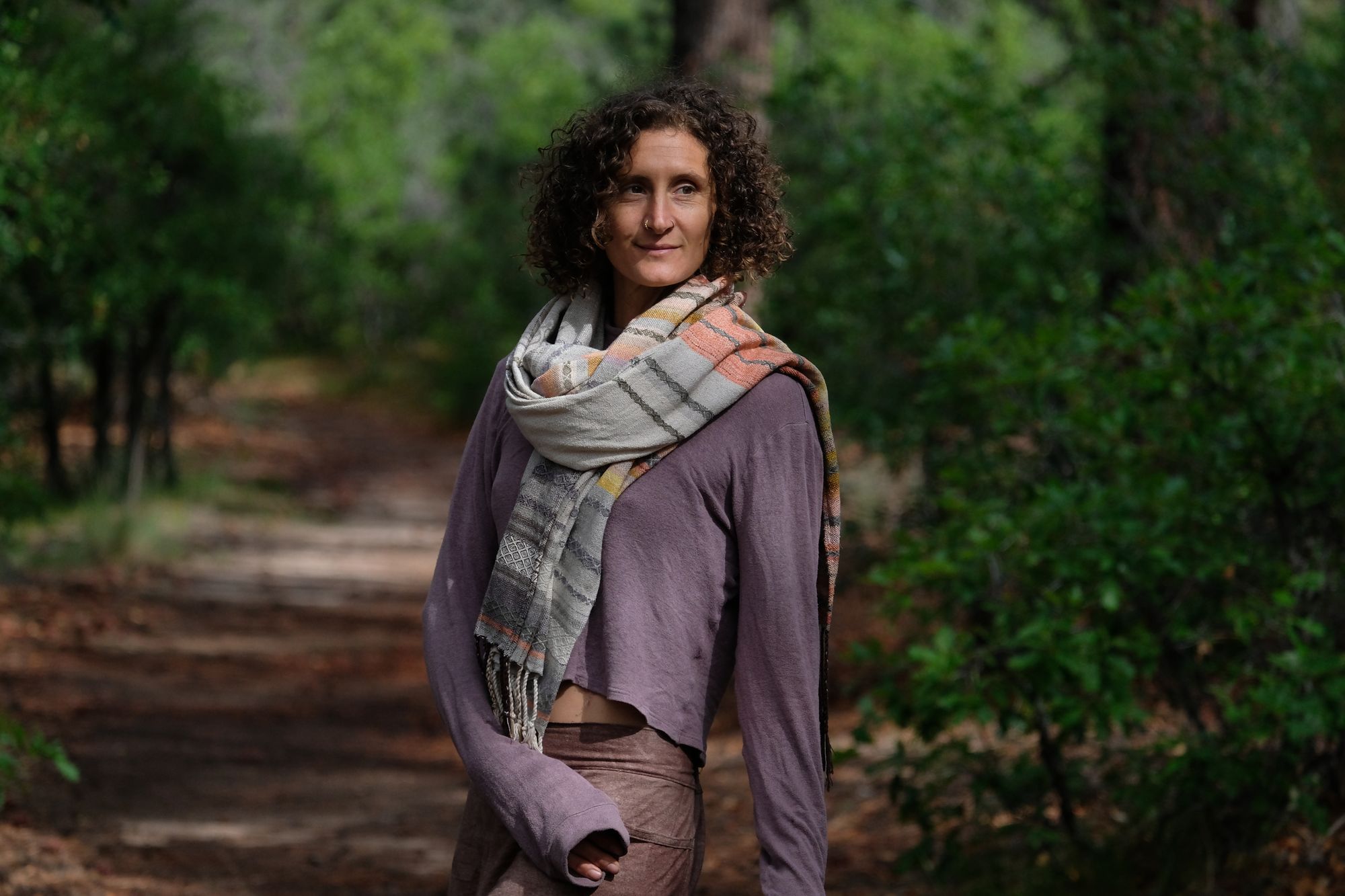 A woman stands on a trail in the forest wearing a handwoven shawl in subtle naturally dyed colors