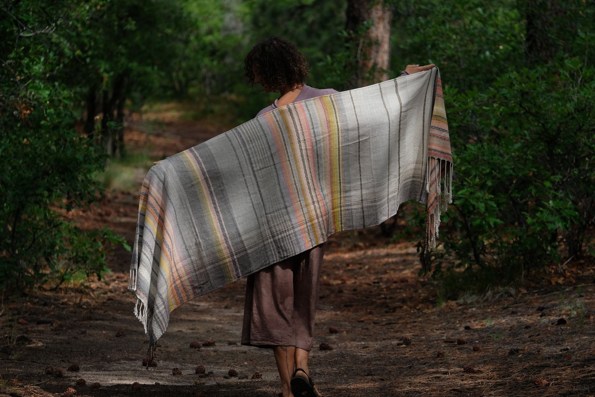 A woman stands on a trail in the forest wearing a handwoven shawl in subtle naturally dyed colors