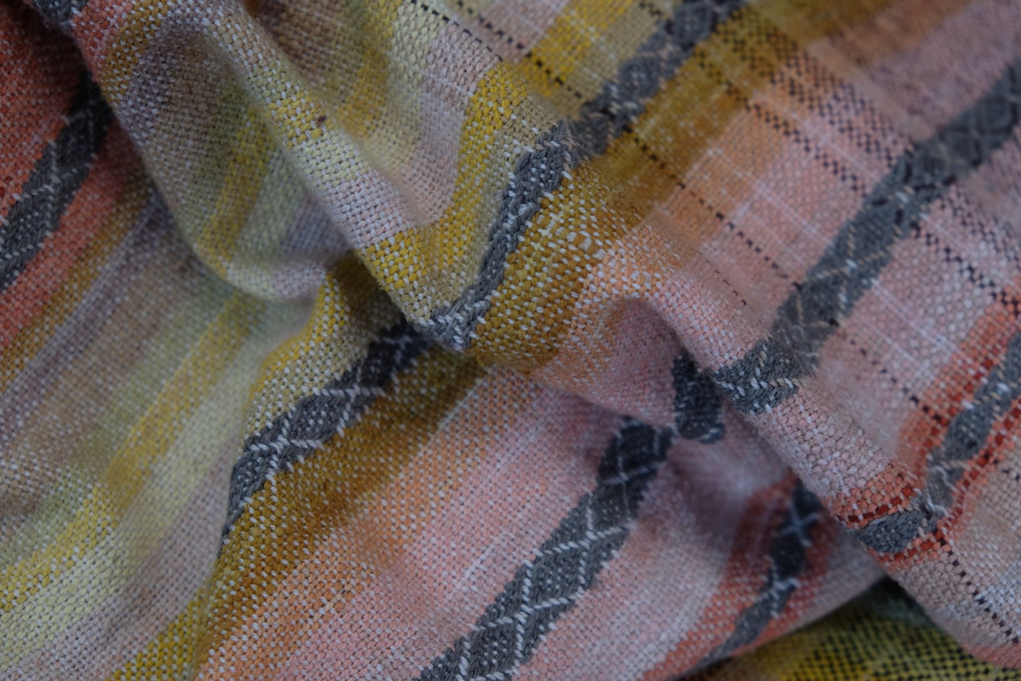 Details of a handwoven shawl in soft but brilliant naturally dyed shades of gray, yellow, pink, salmon, orange, green and purple. The shawl is designed with small stripes of all these colors, and pattern-texture woven details, also of tiny stripes