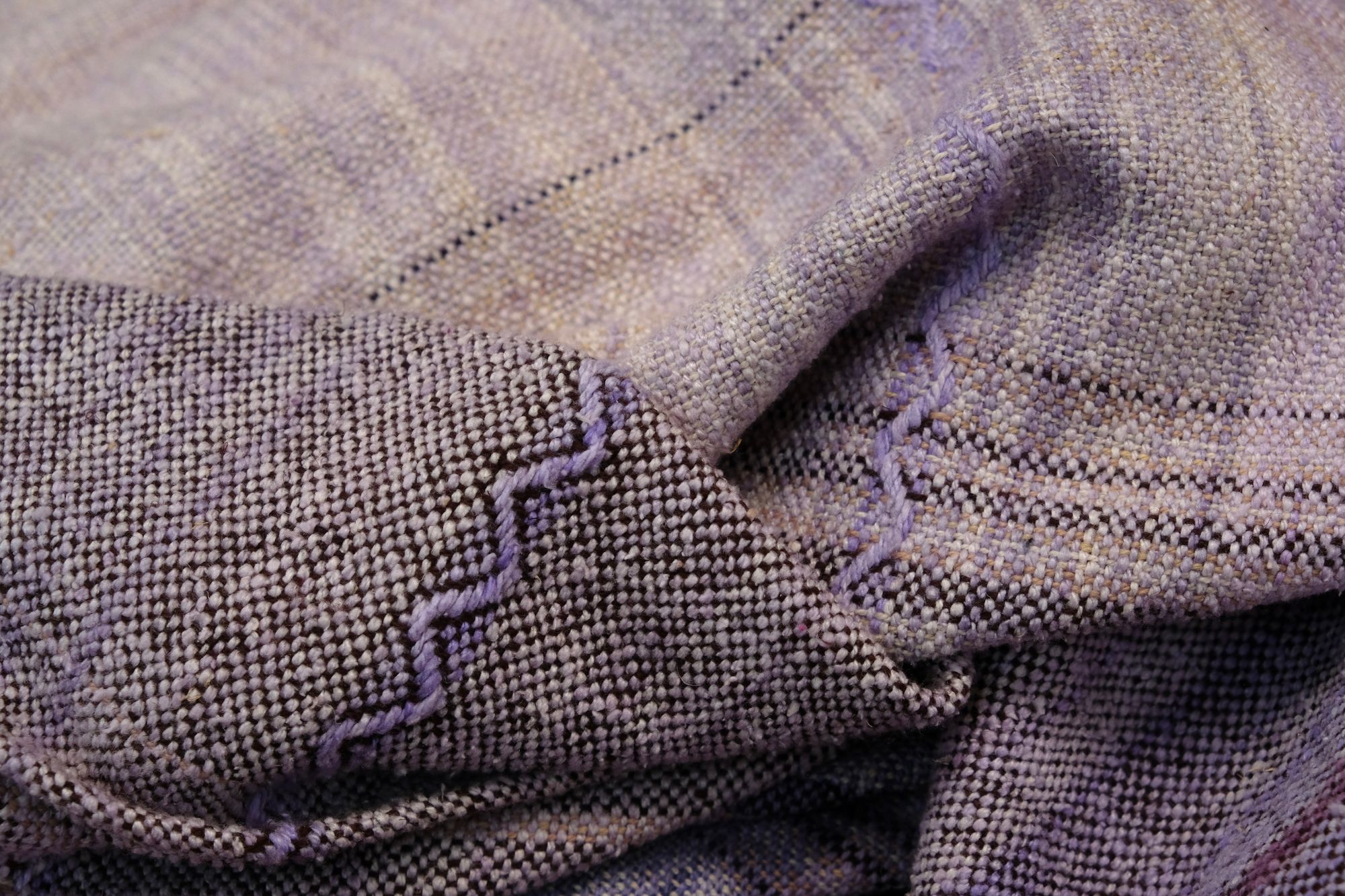 A handwoven length of fabric, woven in every shade of purple with a diamond texture pattern, is laid out on a wooden floor.