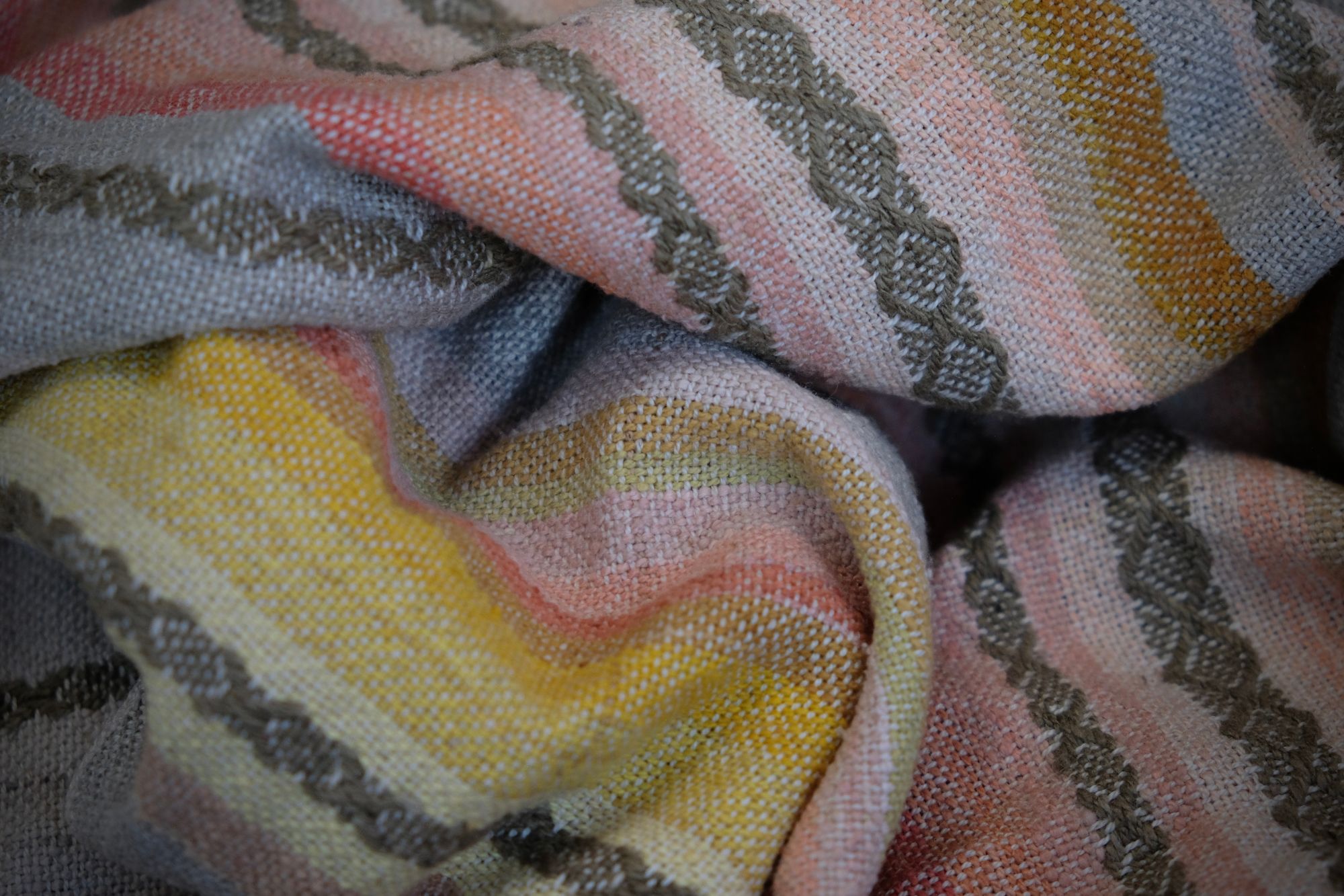Details of a handwoven shawl in soft but brilliant naturally dyed shades of gray, yellow, pink, salmon, orange, green and purple. The shawl is designed with small stripes of all these colors, and pattern-texture woven details, also of tiny stripes