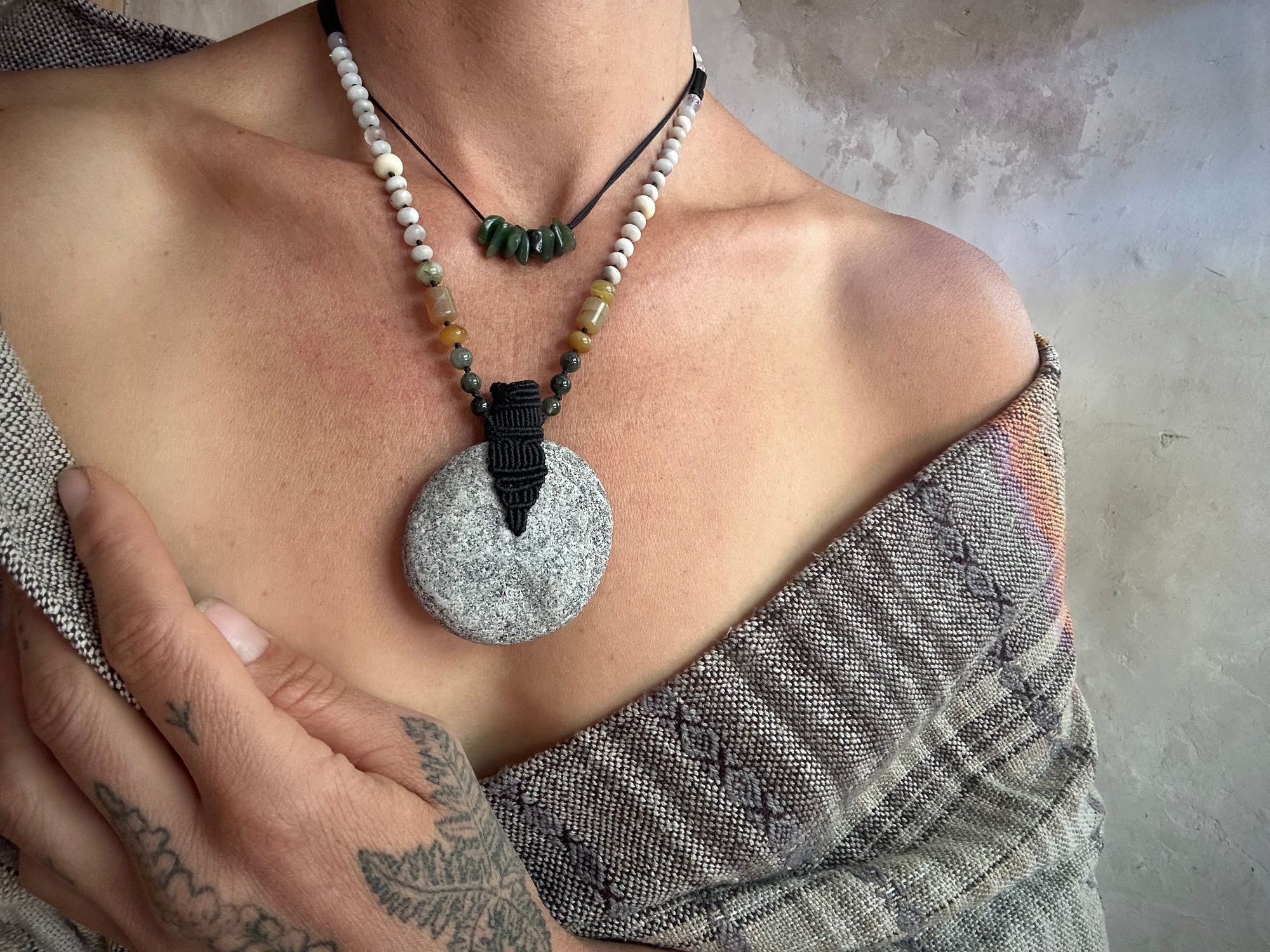 A woman wearing a naturally dyed shawl and tattooed hand wears a stone necklace. At the center the necklace has a granite disc, cradled with a fiber basket. The beads on the necklace are black tourmaline, green rutile quartz spheres, and discs of white jasper and yellow Peruvian opal