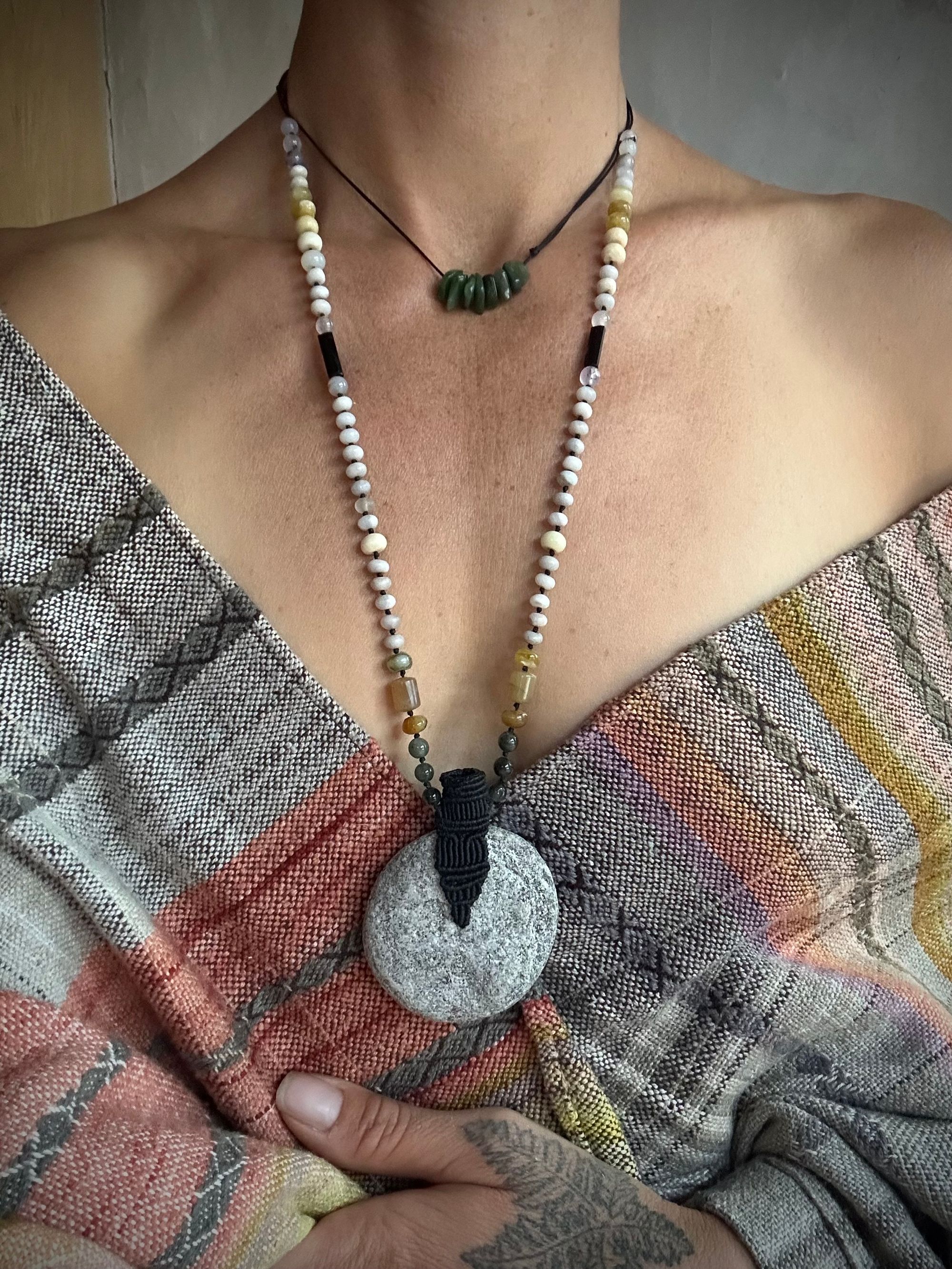 A woman wearing a naturally dyed shawl and tattooed hand wears a stone necklace. At the center the necklace has a granite disc, cradled with a fiber basket. The beads on the necklace are black tourmaline, green rutile quartz spheres, and discs of white jasper and yellow Peruvian opal