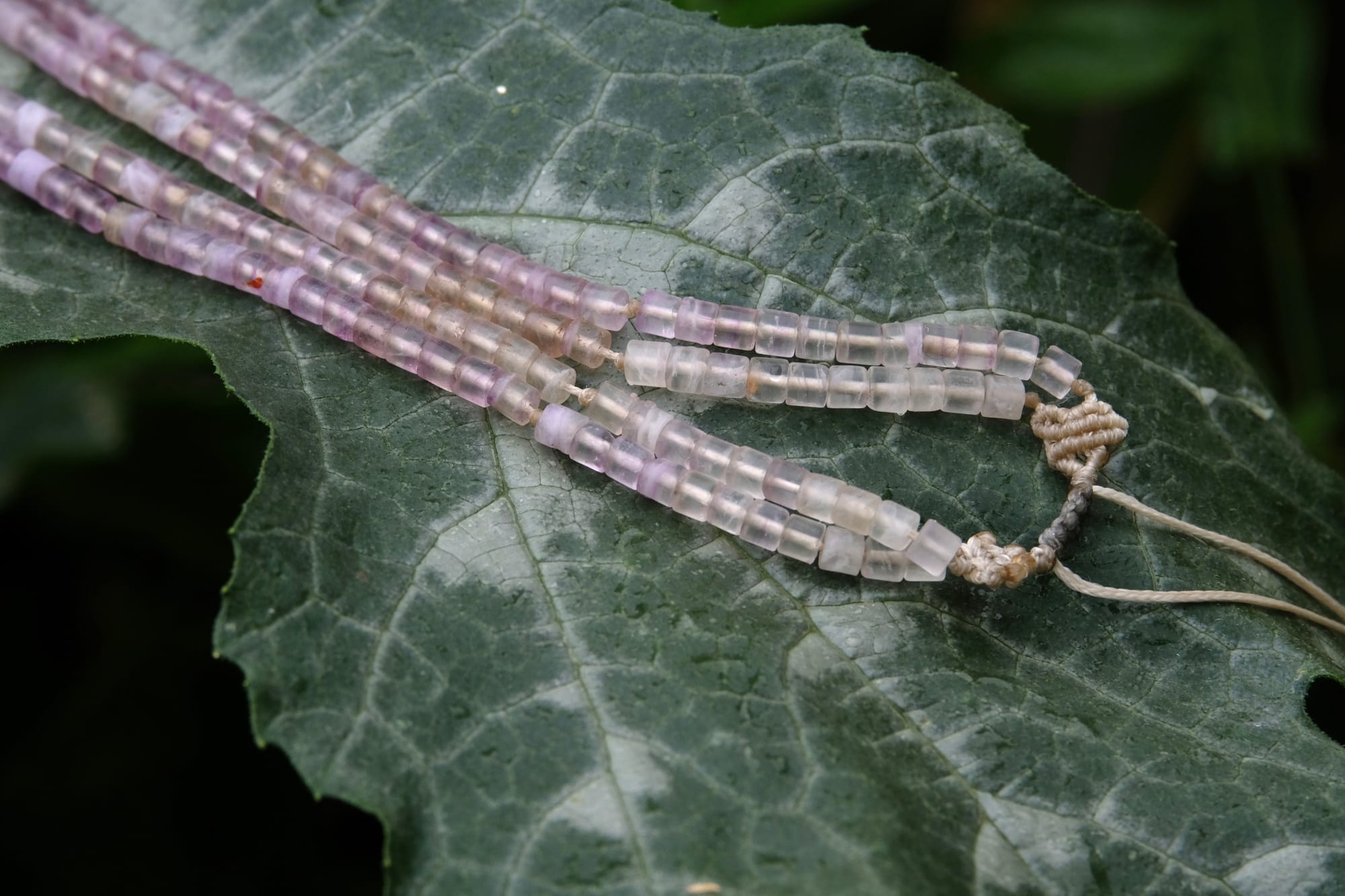 A beaded necklace made of purple fluorite, gray quartz and a clear quartz crystal rests on a large deep green squash leaf