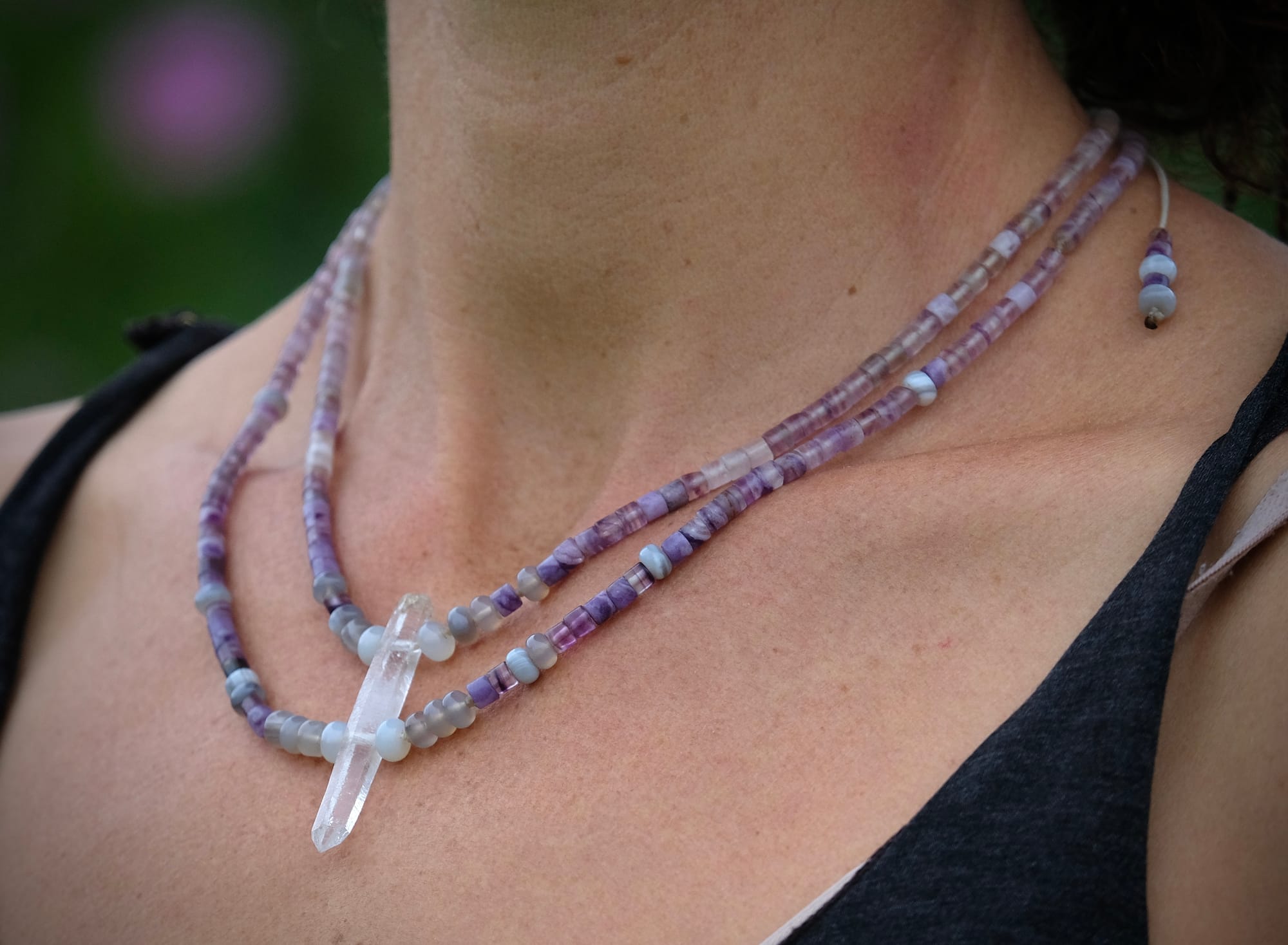 A woman wears a beaded necklace made of purple fluorite, gray quartz and a clear quartz crystal 