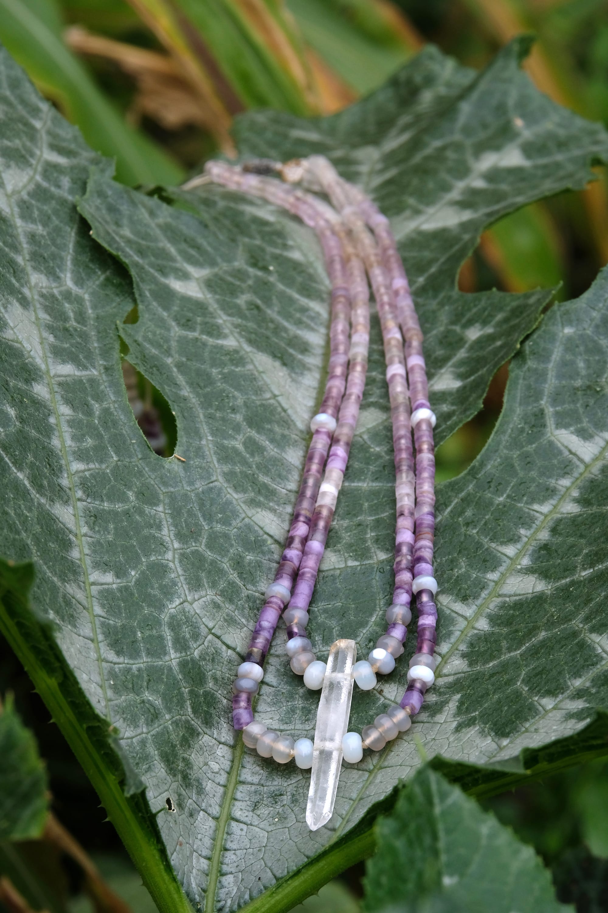 A beaded necklace made of purple fluorite, gray quartz and a clear quartz crystal rests on a large deep green squash leaf