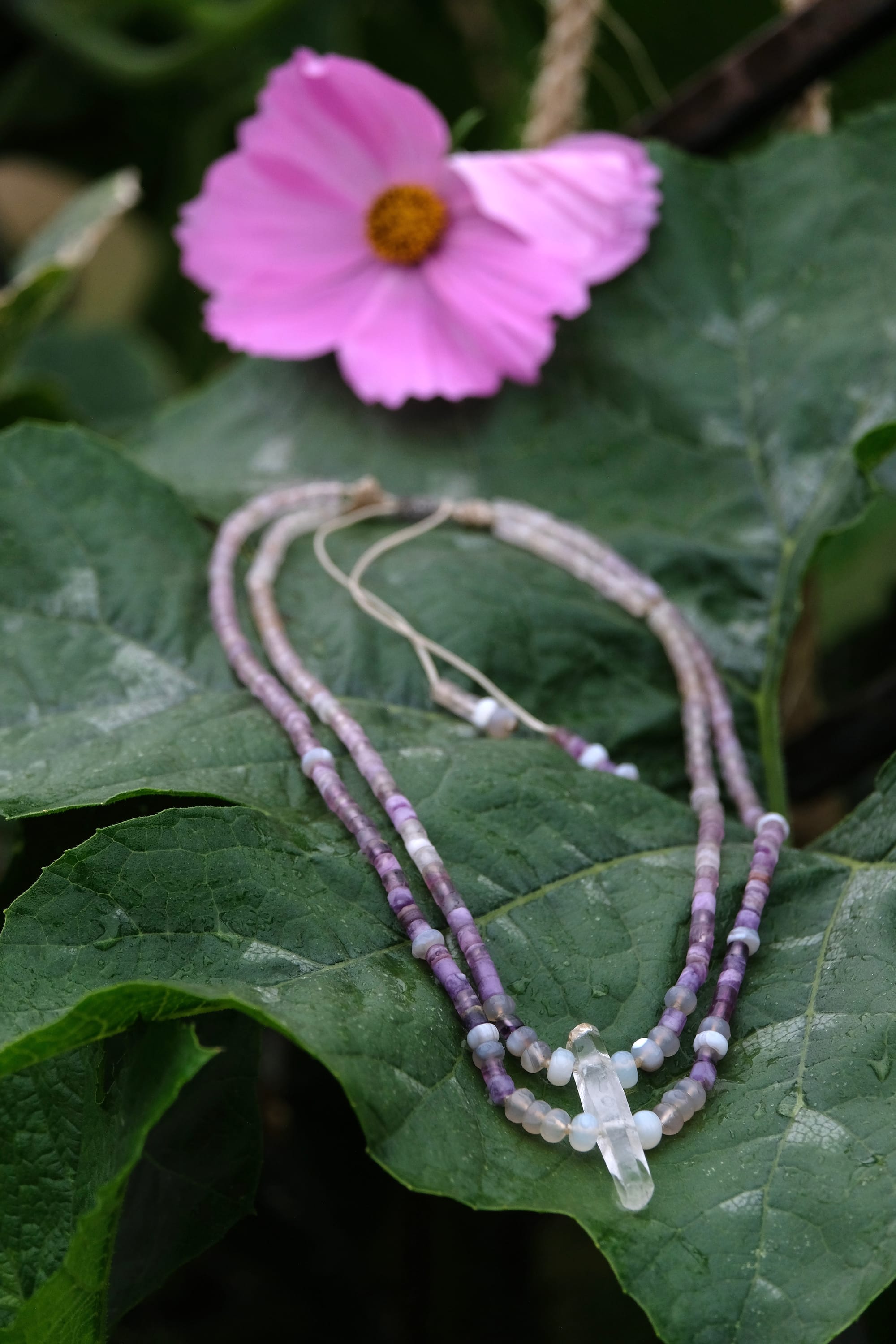 A beaded necklace made of purple fluorite, gray quartz and a clear quartz crystal rests on a large deep green squash leaf