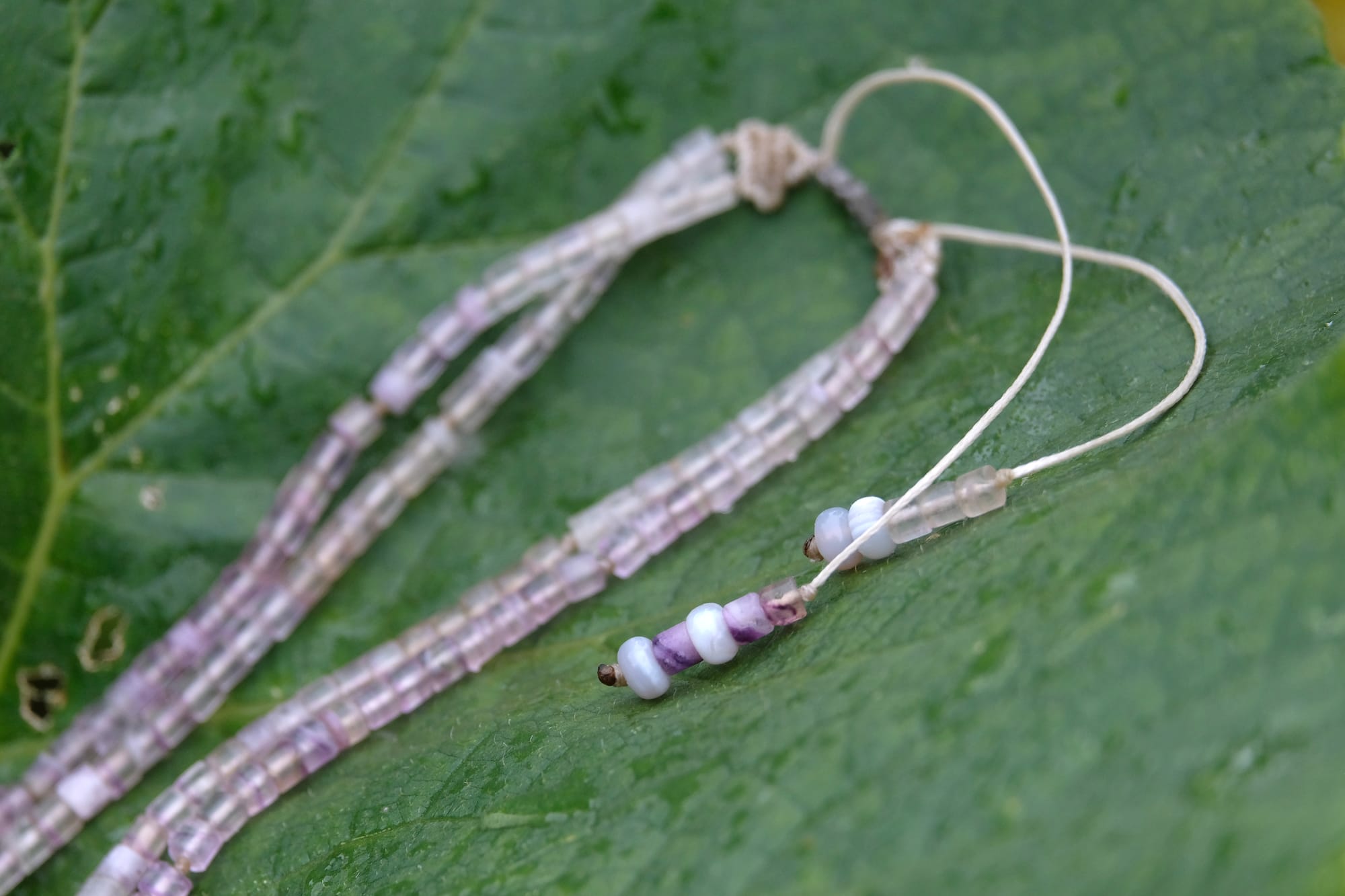 A beaded necklace made of purple fluorite, gray quartz and a clear quartz crystal rests on a large deep green squash leaf