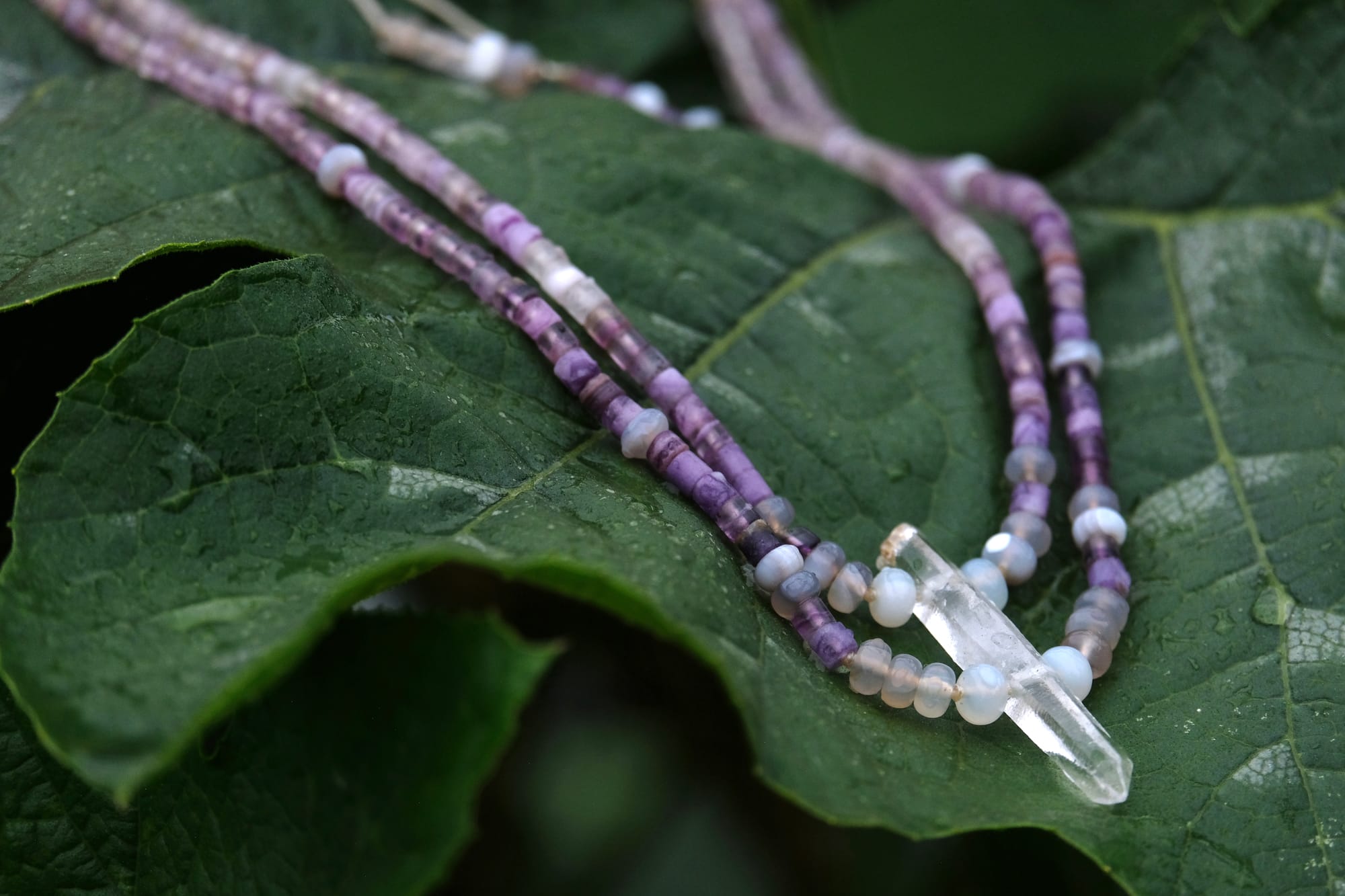 A beaded necklace made of purple fluorite, gray quartz and a clear quartz crystal rests on a large deep green squash leaf
