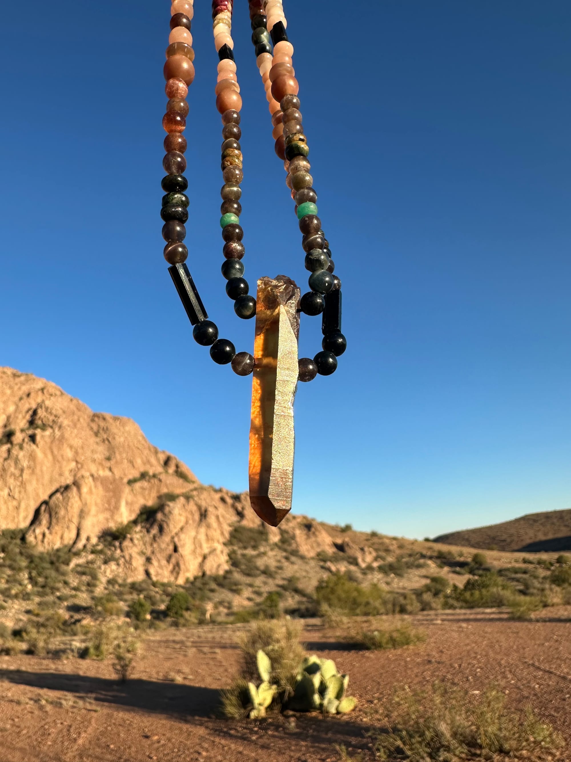 a necklace of luminous semi precious stones in golden citrine, black, dusty pink and deep green hangs against a desert mountain landscape and clear blue sky at sunset