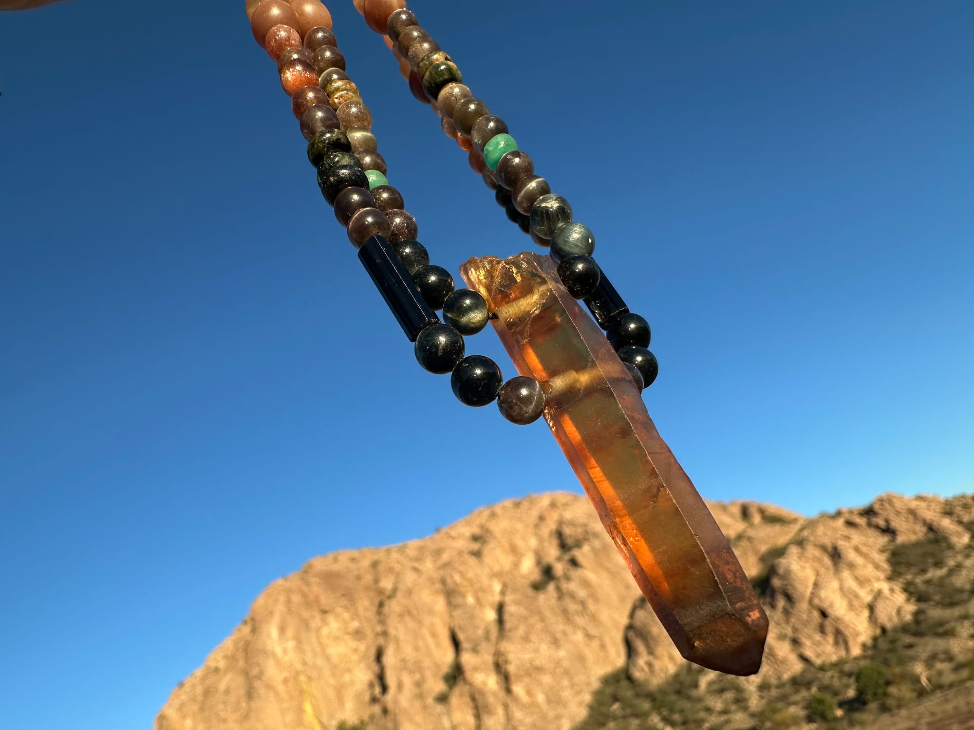 a necklace of luminous semi precious stones in golden citrine, black, dusty pink and deep green hangs against a desert mountain landscape and clear blue sky at sunset