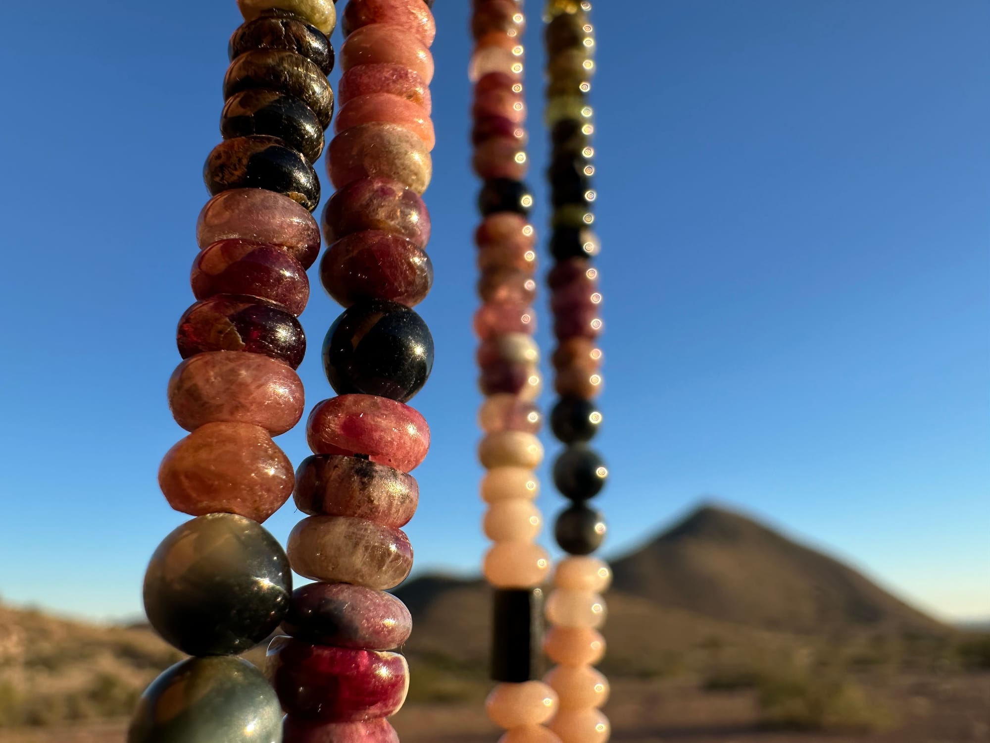 a necklace of luminous semi precious stones in golden citrine, black, dusty pink and deep green hangs against a desert mountain landscape and clear blue sky at sunset