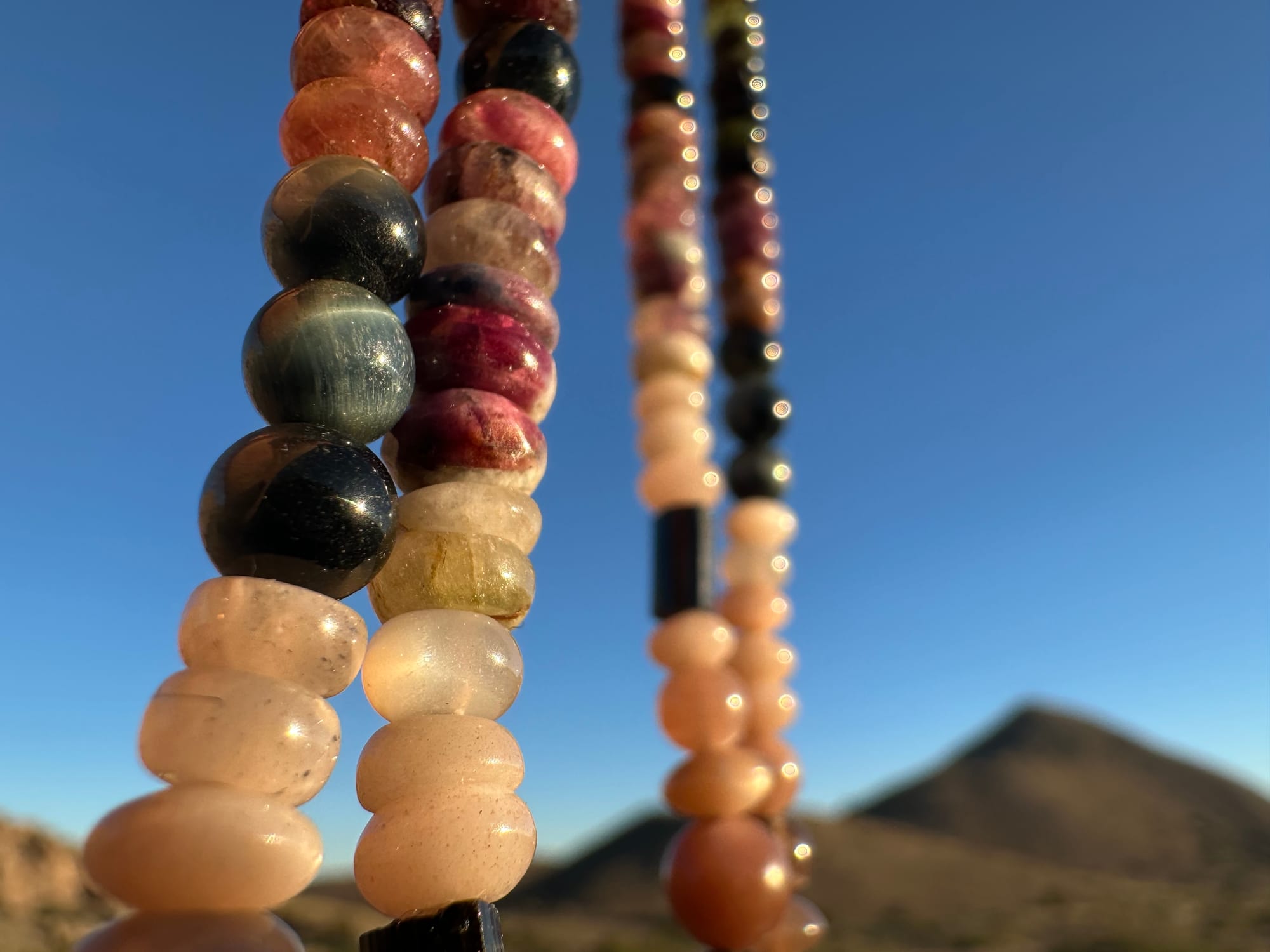 A citrine, cat's eye, tourmaline and emerald necklace by Jeannie Ortiza necklace of luminous semi precious stones in golden citrine, black, dusty pink and deep green hangs against a desert mountain landscape and clear blue sky at sunset