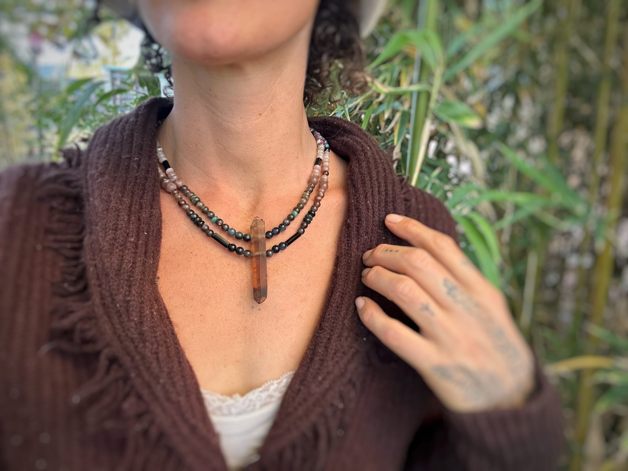 A woman wears a crystal and stone necklace as well as a wooly brown sweater and white wool hat