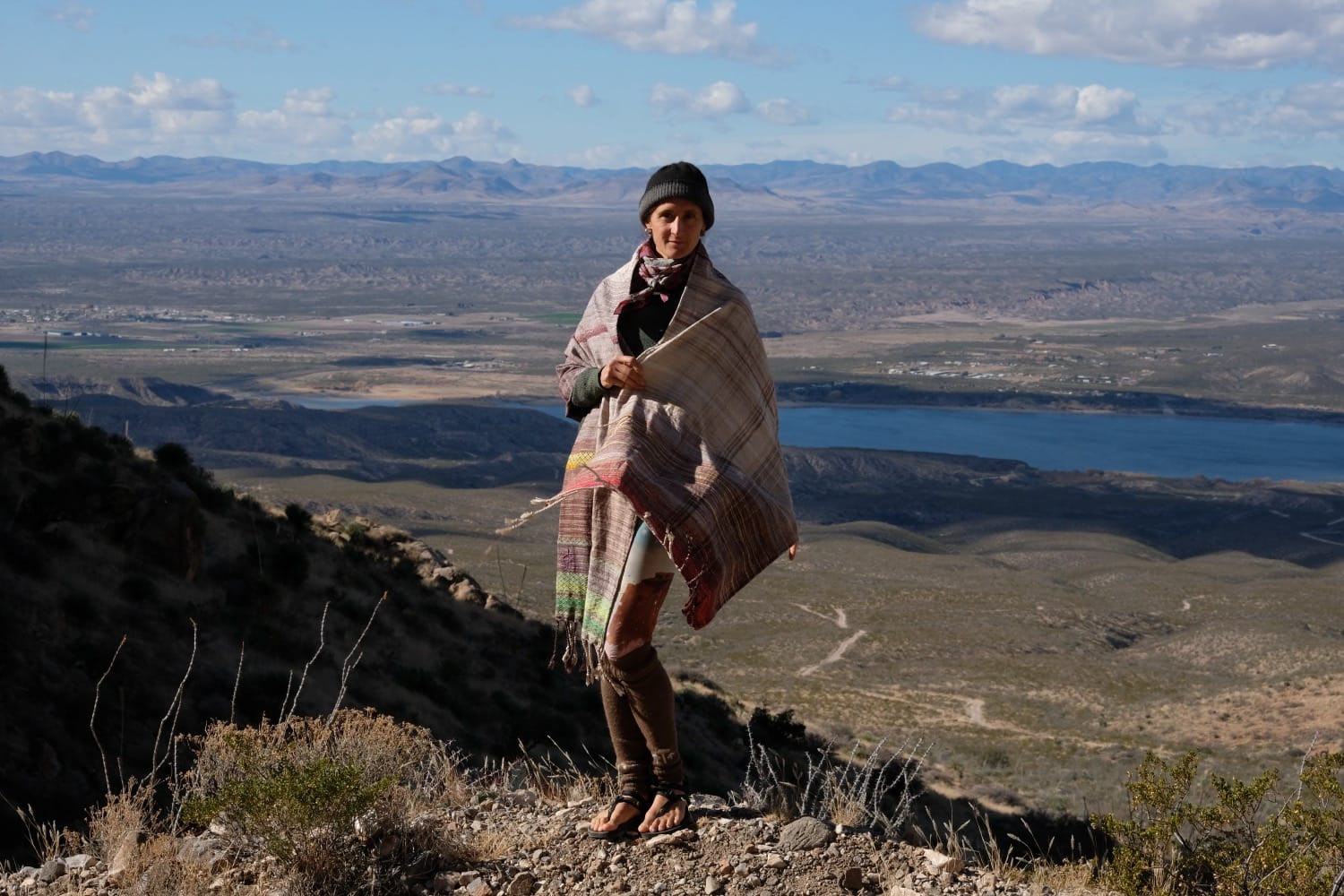 A woman stands on a mountain in a vast desert landscape wearing a handwoven grey, brown, yellow, red and purple shawl