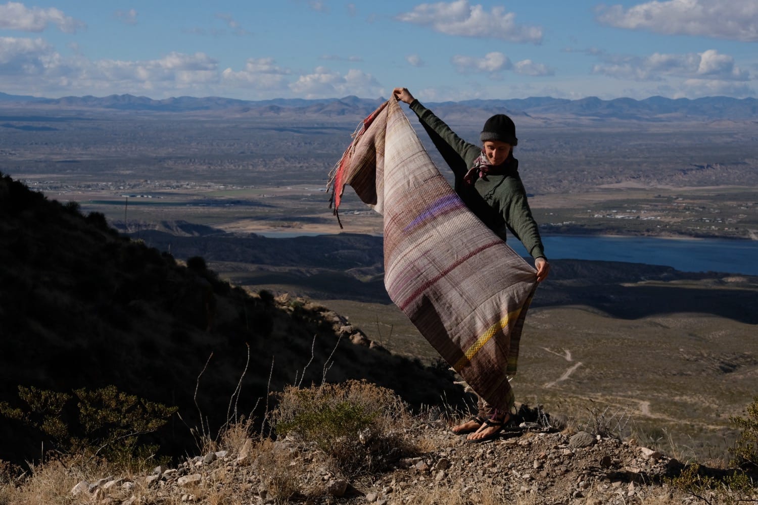 A woman stands on a mountain in a vast desert landscape wearing a handwoven grey, brown, yellow, red and purple shawl