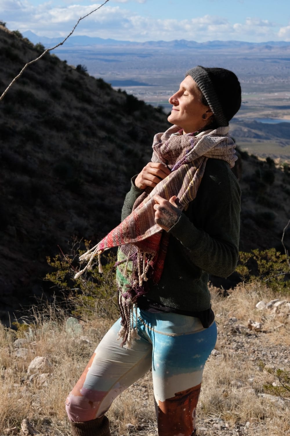 A woman stands on a mountain in a vast desert landscape wearing a handwoven grey, brown, yellow, red and purple shawl