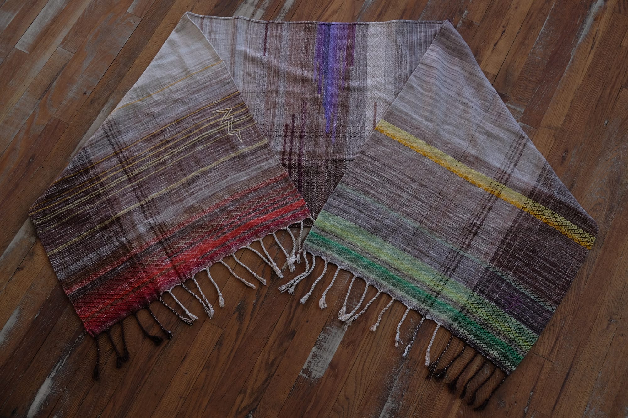 A handwoven shawl with hand dyed raw silk in grey, brown, yellow, purple and red with a lightning bolt detail, on a wooden floor