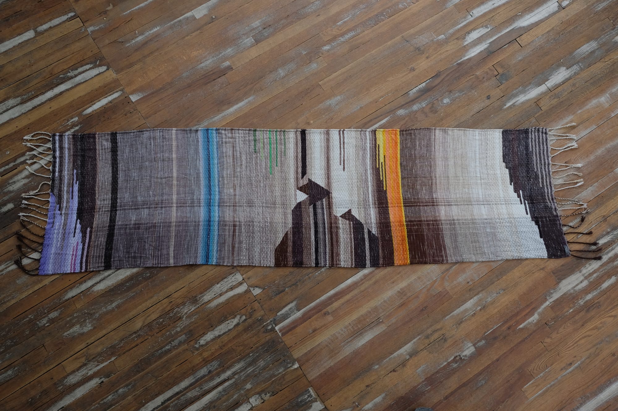 A detail of the geometric tapestry woven designs on a shawl in grayscale, earth-tones and elemental colors of lavender, orange poppy, water and grass. The shawl is laying on a wooden floor